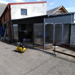 Żółty kompresor Stanley przed częściowo zadaszoną, przeszkloną konstrukcją na posesji z budynkami w tle.