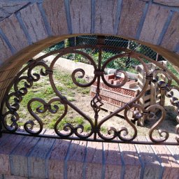 Bartłomiej Lorenc - Dekoracyjny element metalowy w kształcie serc, umieszczony w łukowym otworze z cegły, z widokiem na ogród z siatką ogrodzeniową w tle.