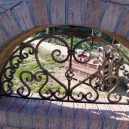 Dekoracyjny element metalowy w kształcie serc, umieszczony w łukowym otworze z cegły, z widokiem na ogród z siatką ogrodzeniową w tle.