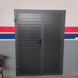 Bartłomiej Lorenc - Zamknięta, dwuskrzydłowa, szara brama garażowa z poziomymi panelami, zamontowana w szarej ścianie z czerwono-niebieskim pasem dekoracyjnym.