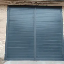 Zamknięta, dwuczęściowa, szara brama rolowana na tle ceglanego budynku z widoczną instalacją elektryczną po lewej stronie.