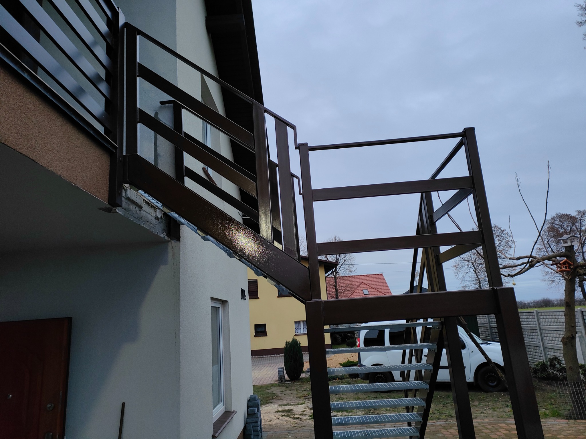 Zewnętrzne metalowe schody z balustradą, prowadzące do balkonu domu jednorodzinnego, widok z dołu, szare niebo w tle.