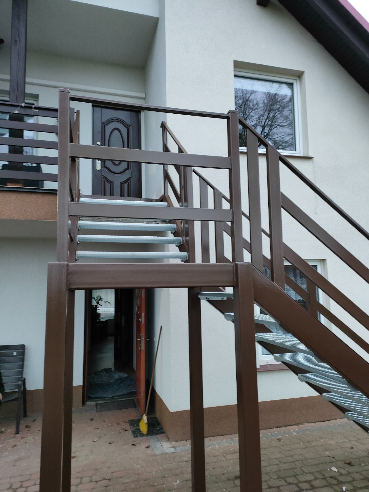 Zewnętrzne schody metalowe z brązową balustradą i ażurowymi stopniami prowadzące do drzwi wejściowych na piętrze budynku.
