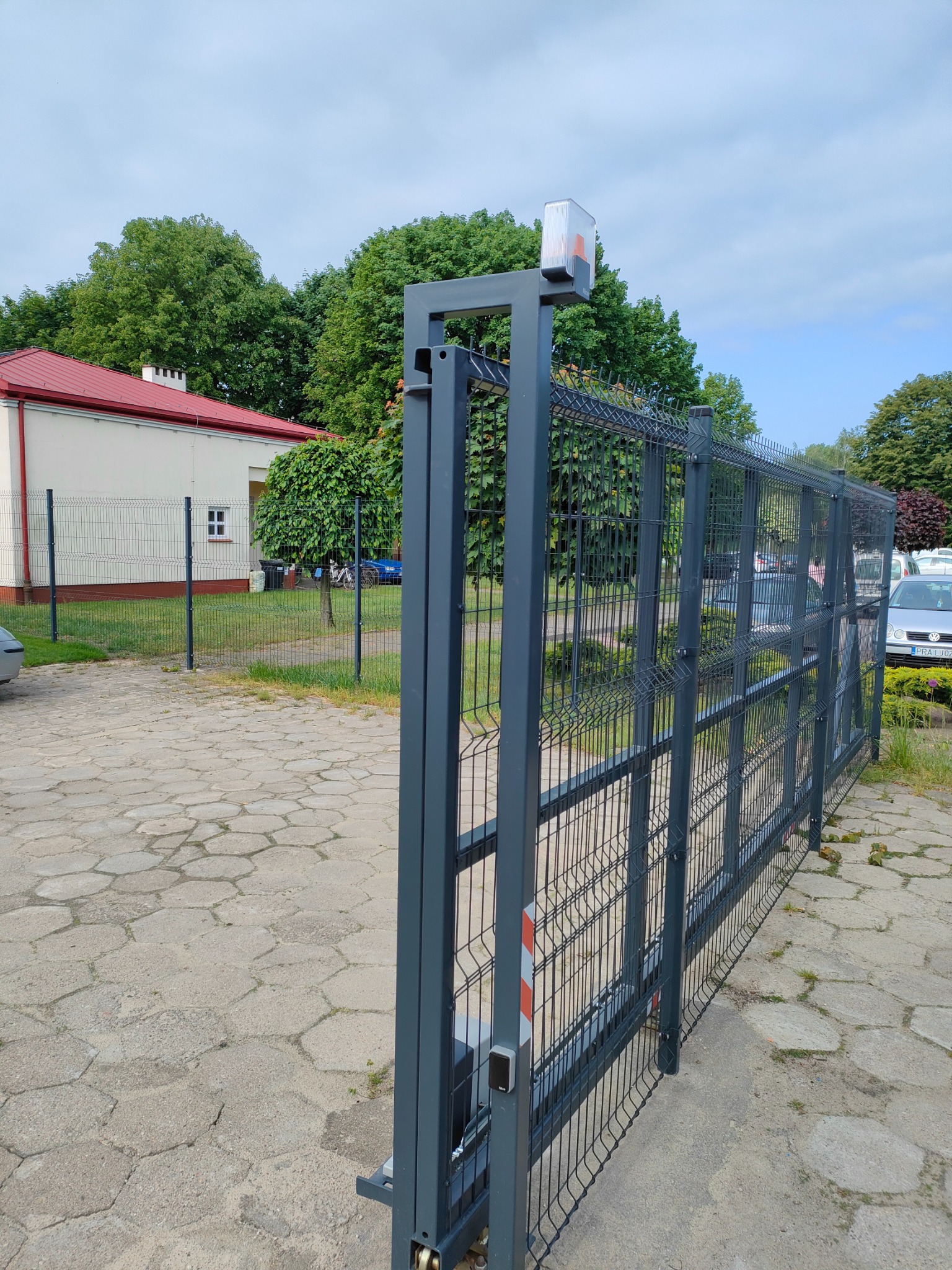 Nowoczesna, przesuwna brama wjazdowa z szarej siatki panelowej z automatyką, widoczna na tle budynku z czerwonym dachem i zielenią.