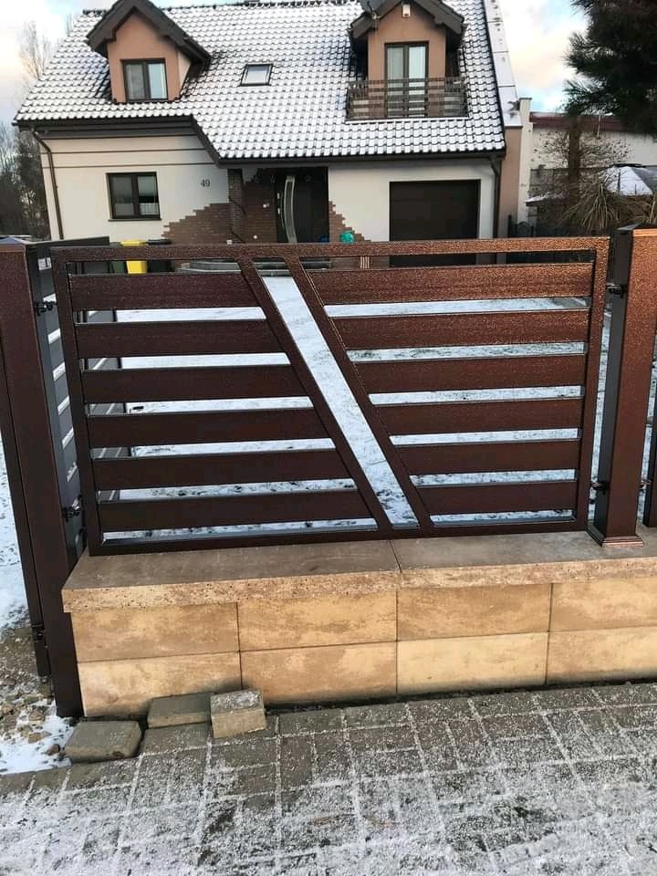 Nowoczesne, poziome ogrodzenie metalowe w kolorze brązowym z diagonalnym akcentem, zamontowane na niskim murku z jasnego kamienia, na tle ośnieżonego chodnika i domu.