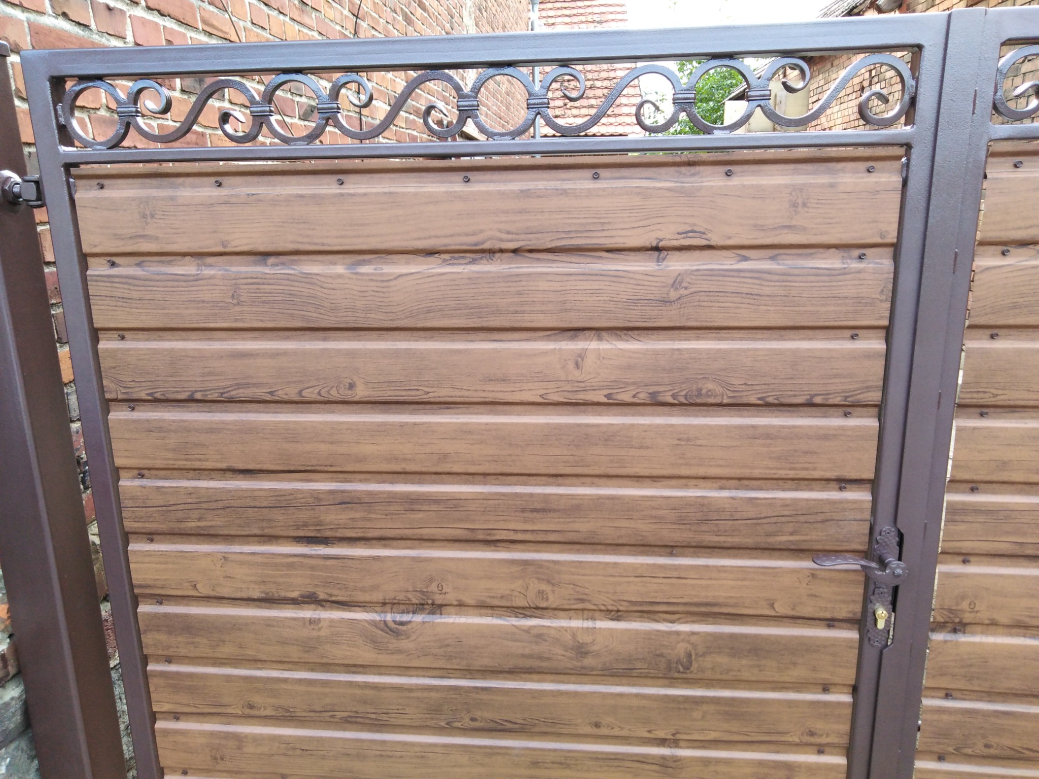 Brązowa brama z panelami imitującymi drewno i ozdobnym metalowym wykończeniem u góry, widoczny zamek i klamka.