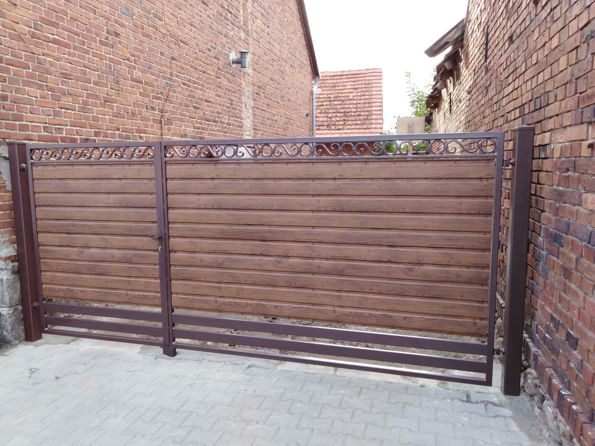 Nowoczesna brama wjazdowa z poziomych paneli imitujących drewno, z metalową ramą i ozdobnymi elementami na górze, na tle ceglanego budynku.