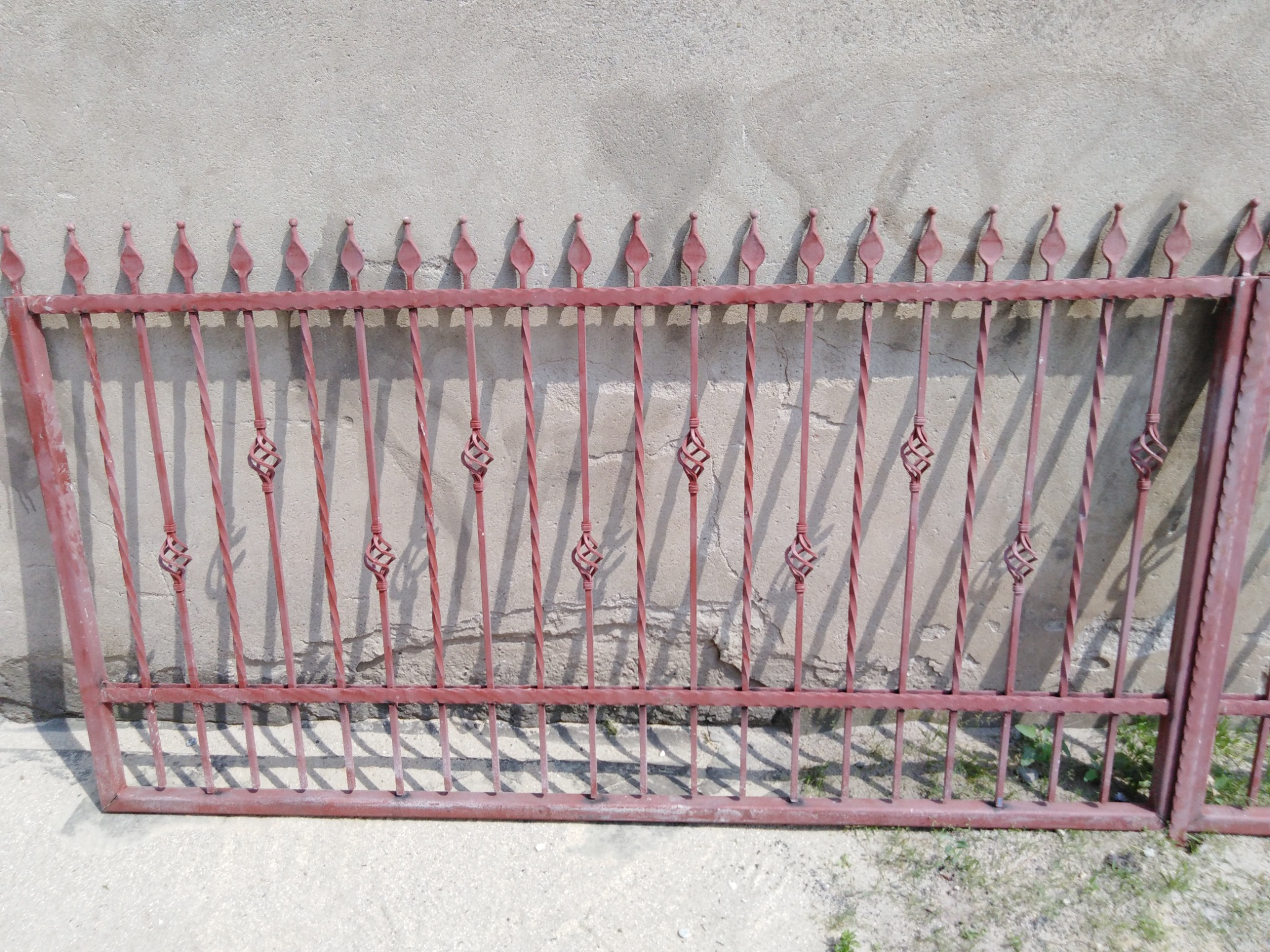 Fragment metalowego ogrodzenia w kolorze ceglastym, leżącego na ziemi przed szarą ścianą, z widocznymi spawami i ozdobnymi elementami w kształcie koszyczków na pionowych prętach.