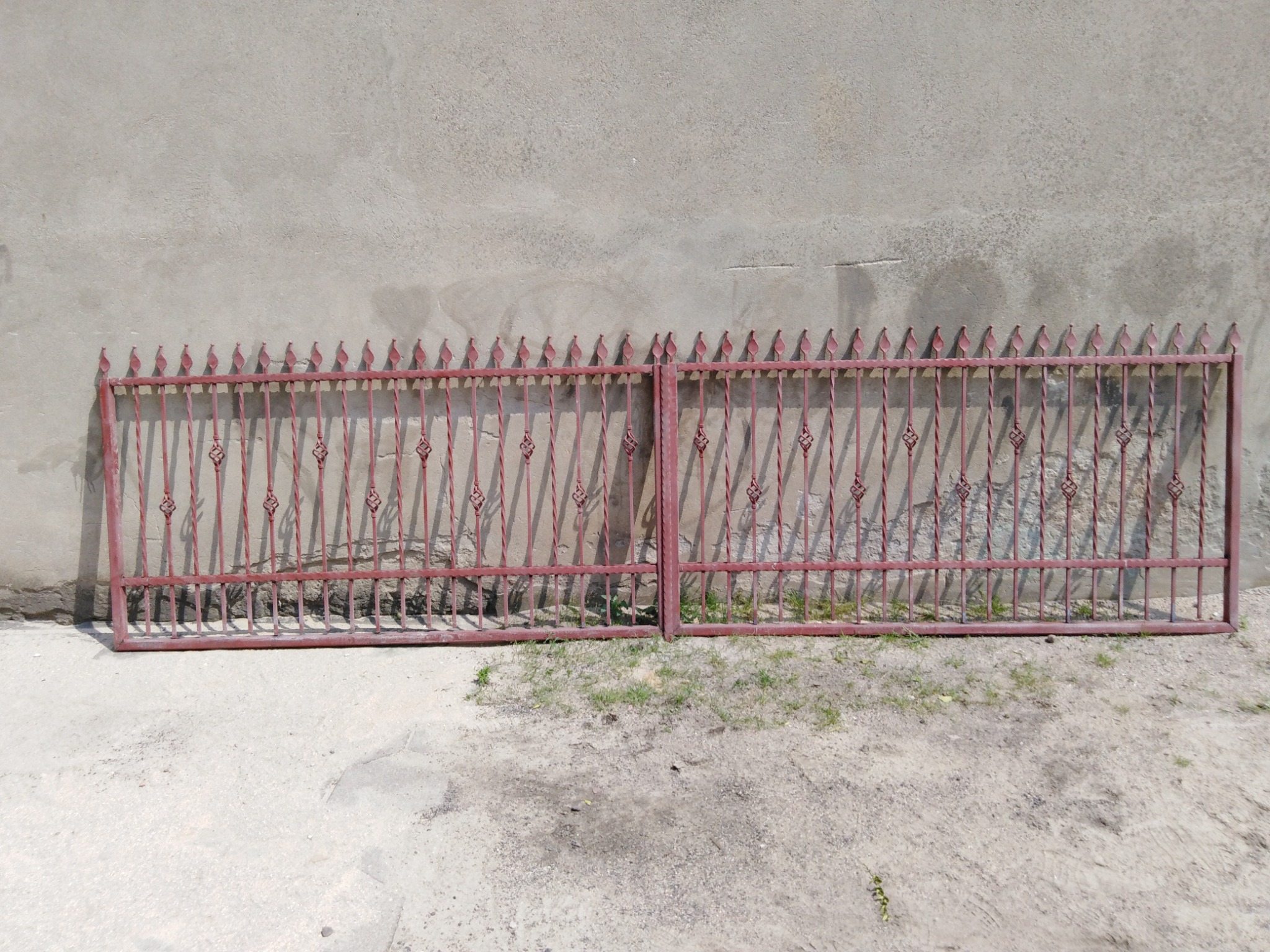 Dwie połówki metalowej bramy w kolorze bordowym, z dekoracyjnymi elementami w kształcie liści i ostrymi zakończeniami, leżące na ziemi przed szarą ścianą.