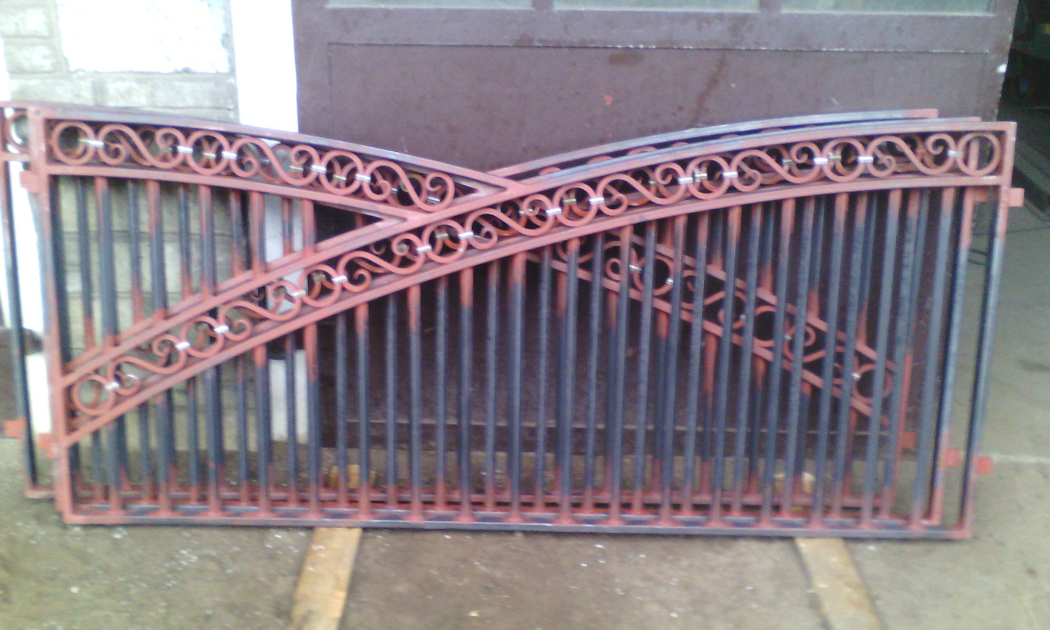 Dwie metalowe bramy wjazdowe z dekoracyjnymi, zakrzywionymi elementami, pomalowane w kolorze ceglastym i czarnym, leżące na ziemi przed budynkiem.