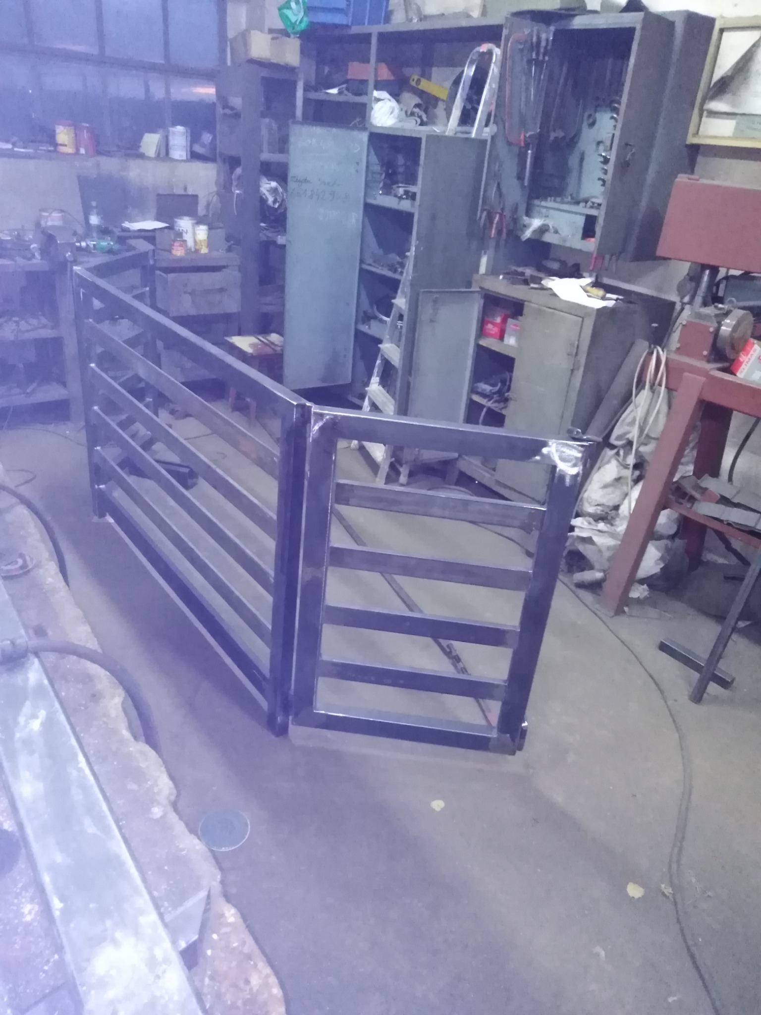 Surowa, spawana konstrukcja metalowa, przypominająca fragment ogrodzenia lub bramki, widoczna w warsztacie z narzędziami i szafkami w tle.