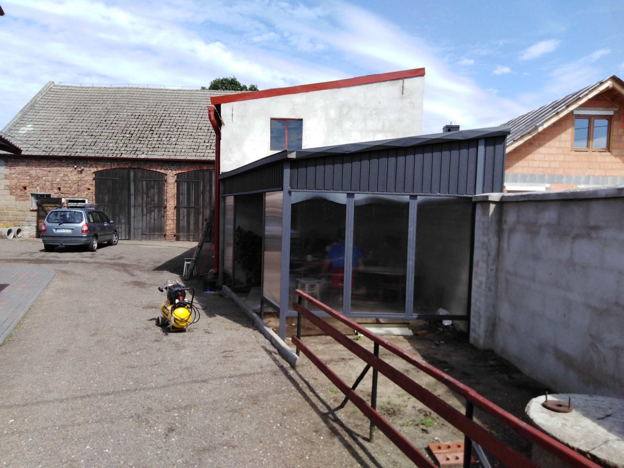 Szara, przeszklona konstrukcja z metalowym dachem, prawdopodobnie altana lub wiata, w trakcie montażu na posesji w Miejskiej Górce. W tle budynki gospodarcze i kompresor. Widoczny fragment...