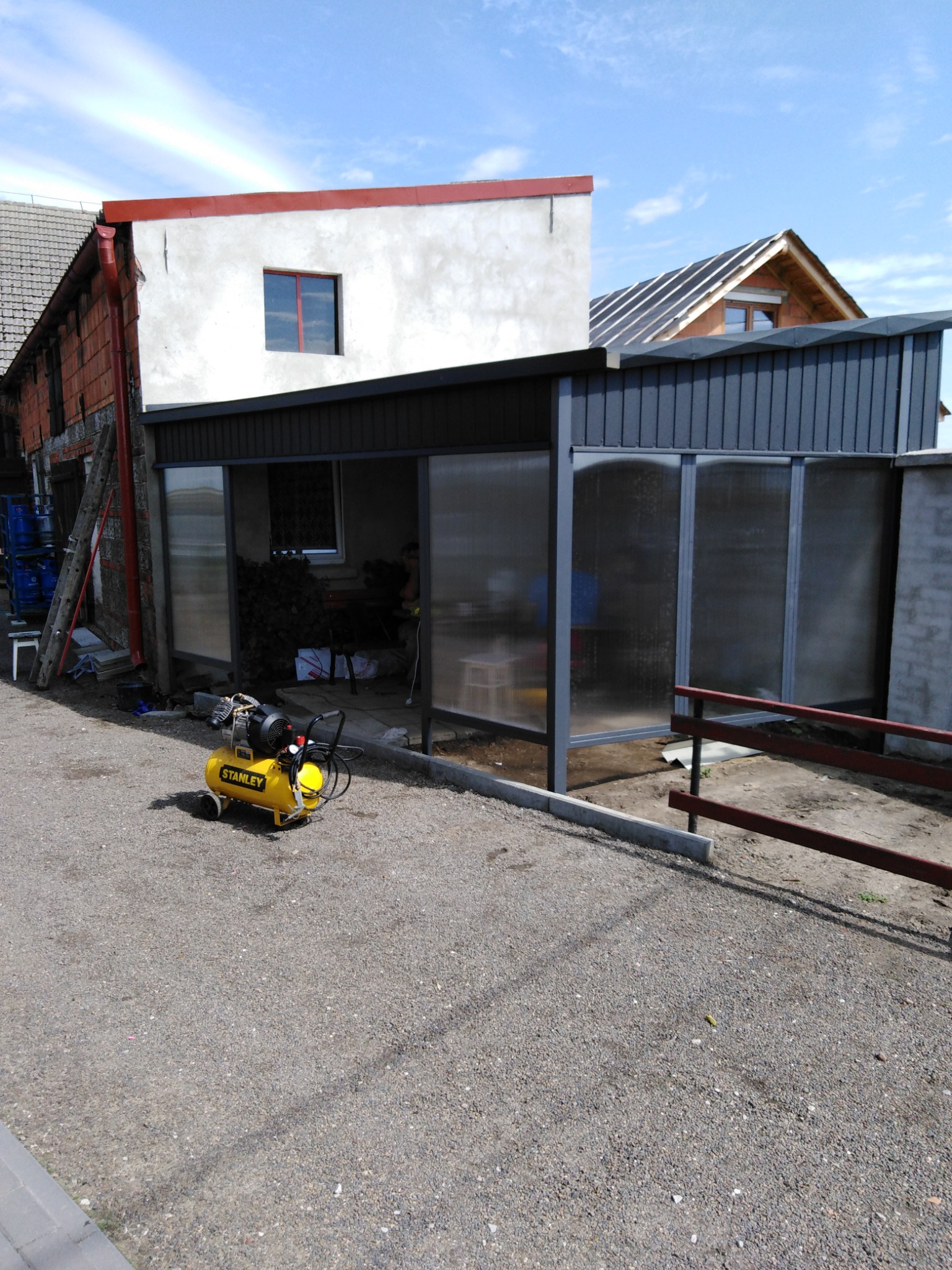 Żółty kompresor Stanley przed częściowo zadaszoną, przeszkloną konstrukcją na posesji z budynkami w tle.