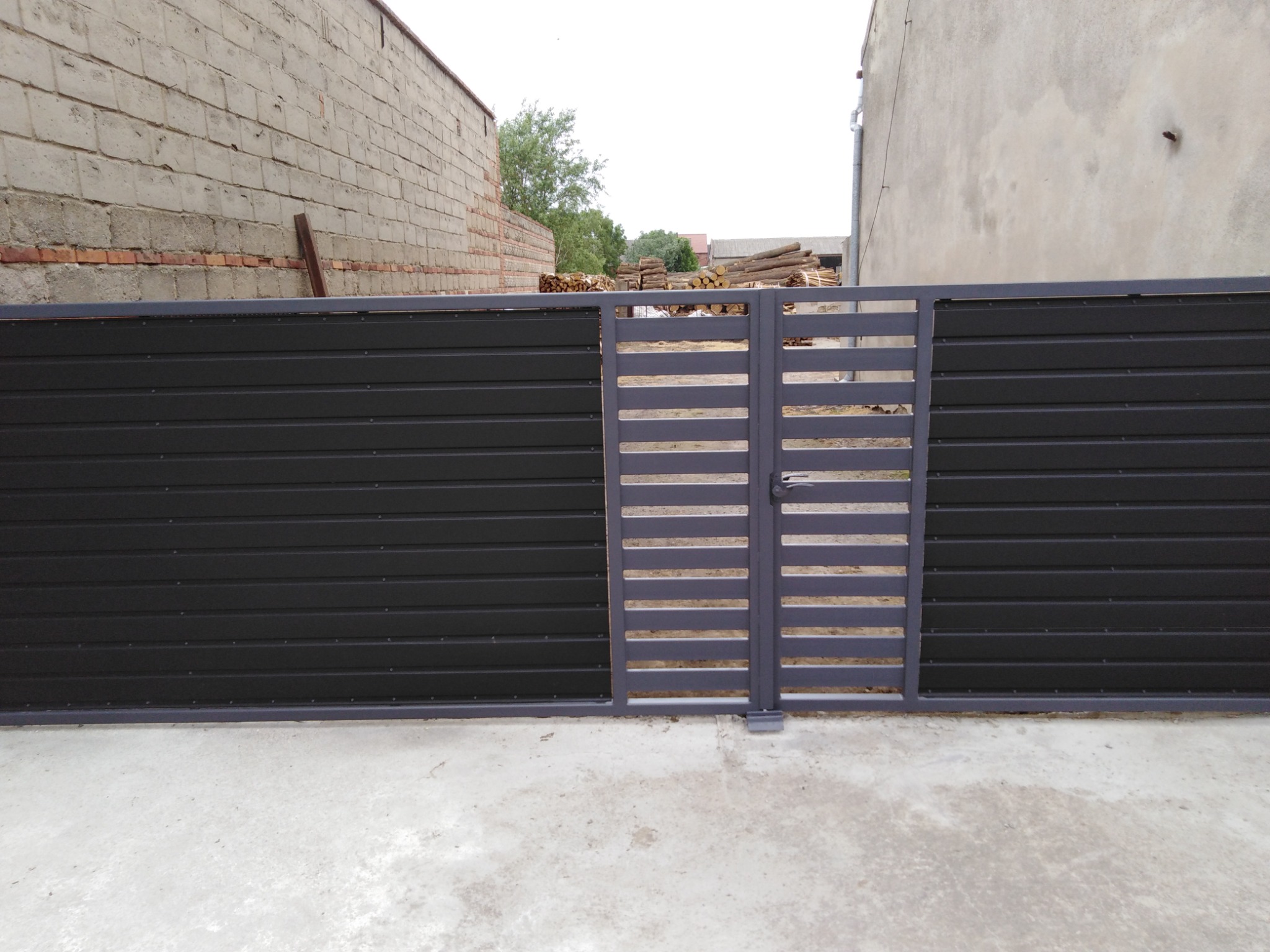 Nowoczesna brama wjazdowa z poziomych paneli w kolorze grafitowym i szarym, na tle betonowego placu i budynków gospodarczych.
