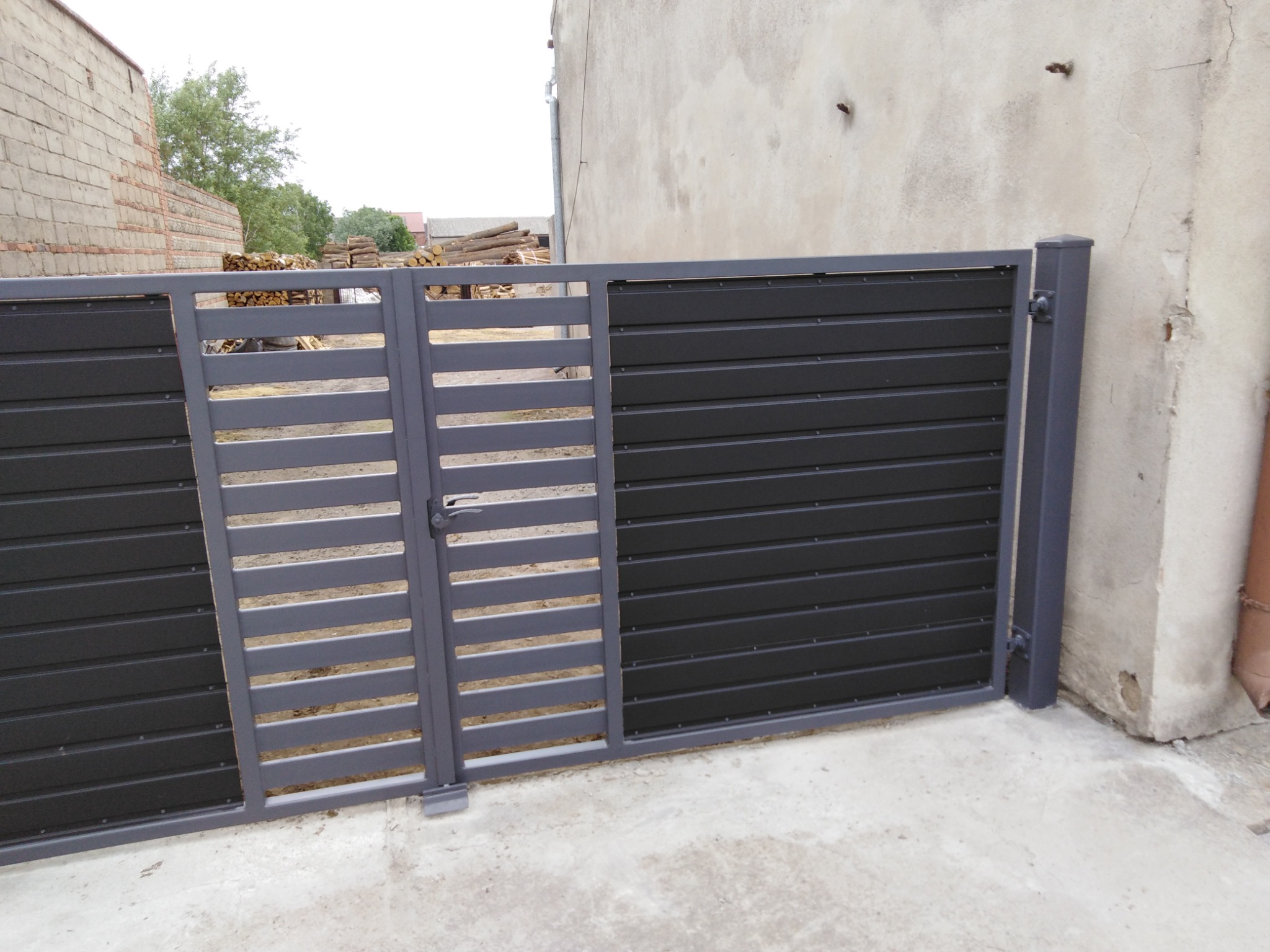 Nowoczesna brama wjazdowa z elementami panelowymi w kolorze szaro-czarnym, widoczna na tle sterty drewna i budynku gospodarczego.