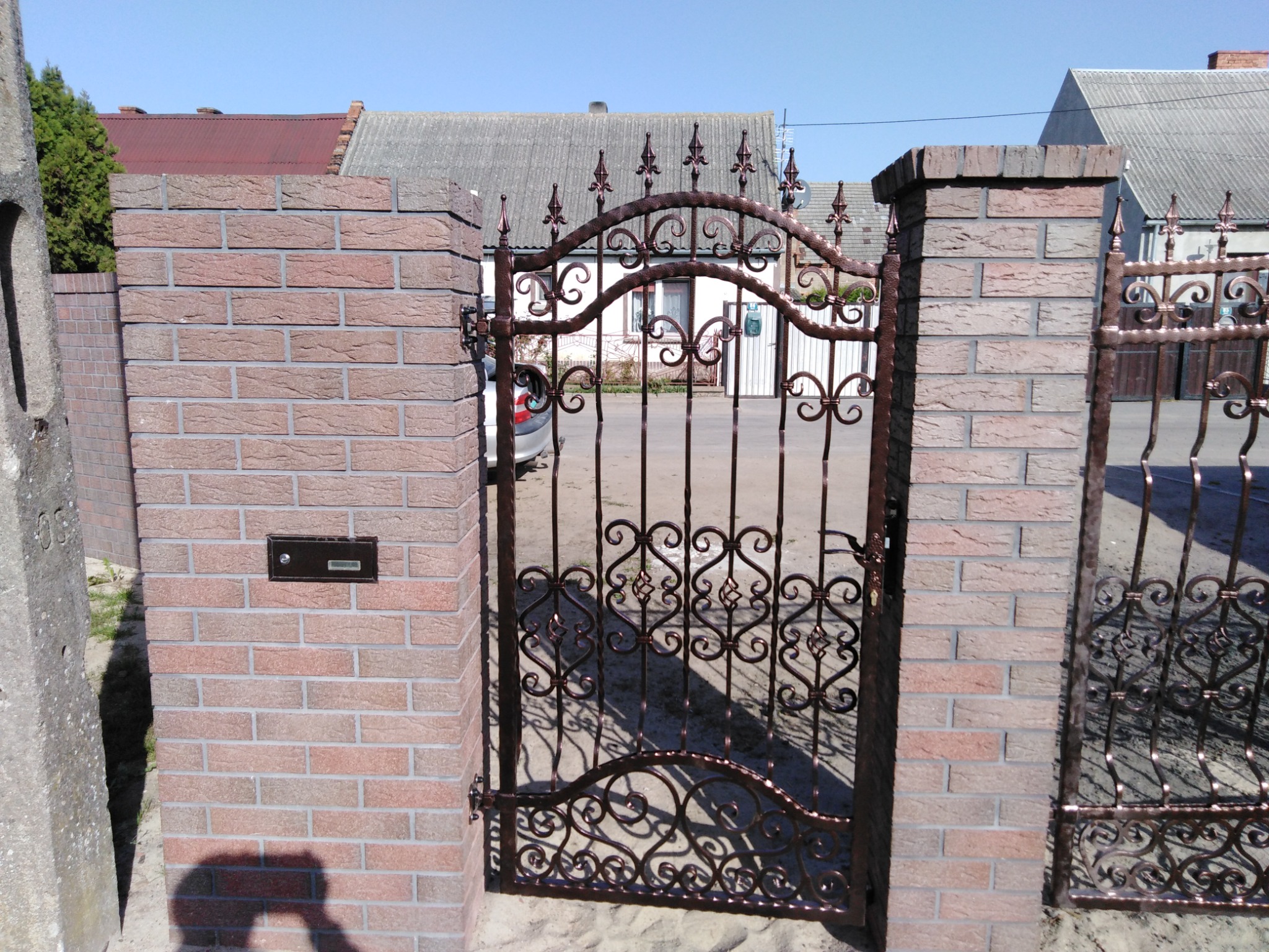 Ozdobna, metalowa brama wjazdowa z dekoracyjnymi elementami, osadzona w murze z cegły klinkierowej z zamontowaną skrzynką na listy.