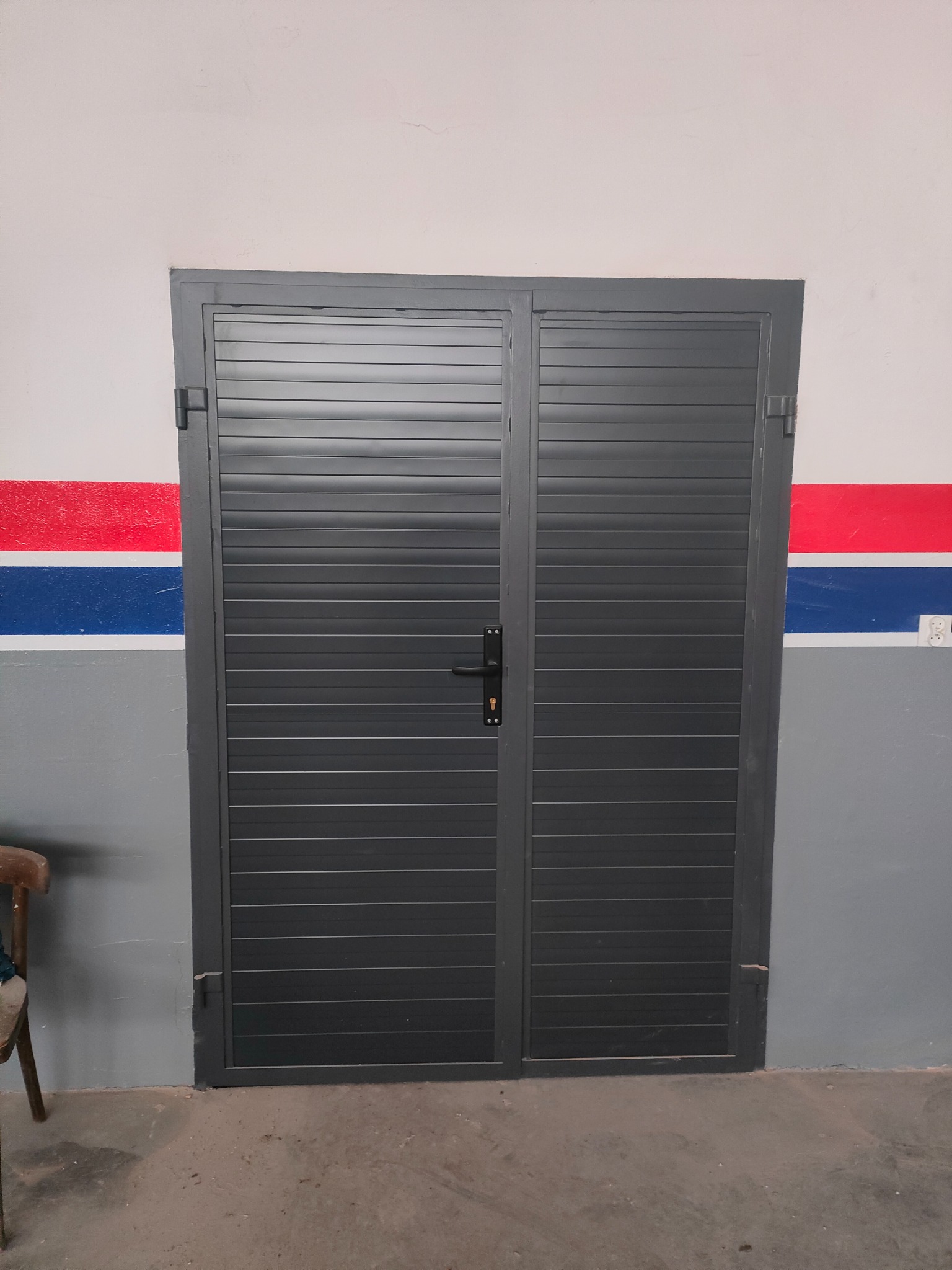 Zamknięta, dwuskrzydłowa, szara brama garażowa z poziomymi panelami, zamontowana w szarej ścianie z czerwono-niebieskim pasem dekoracyjnym.