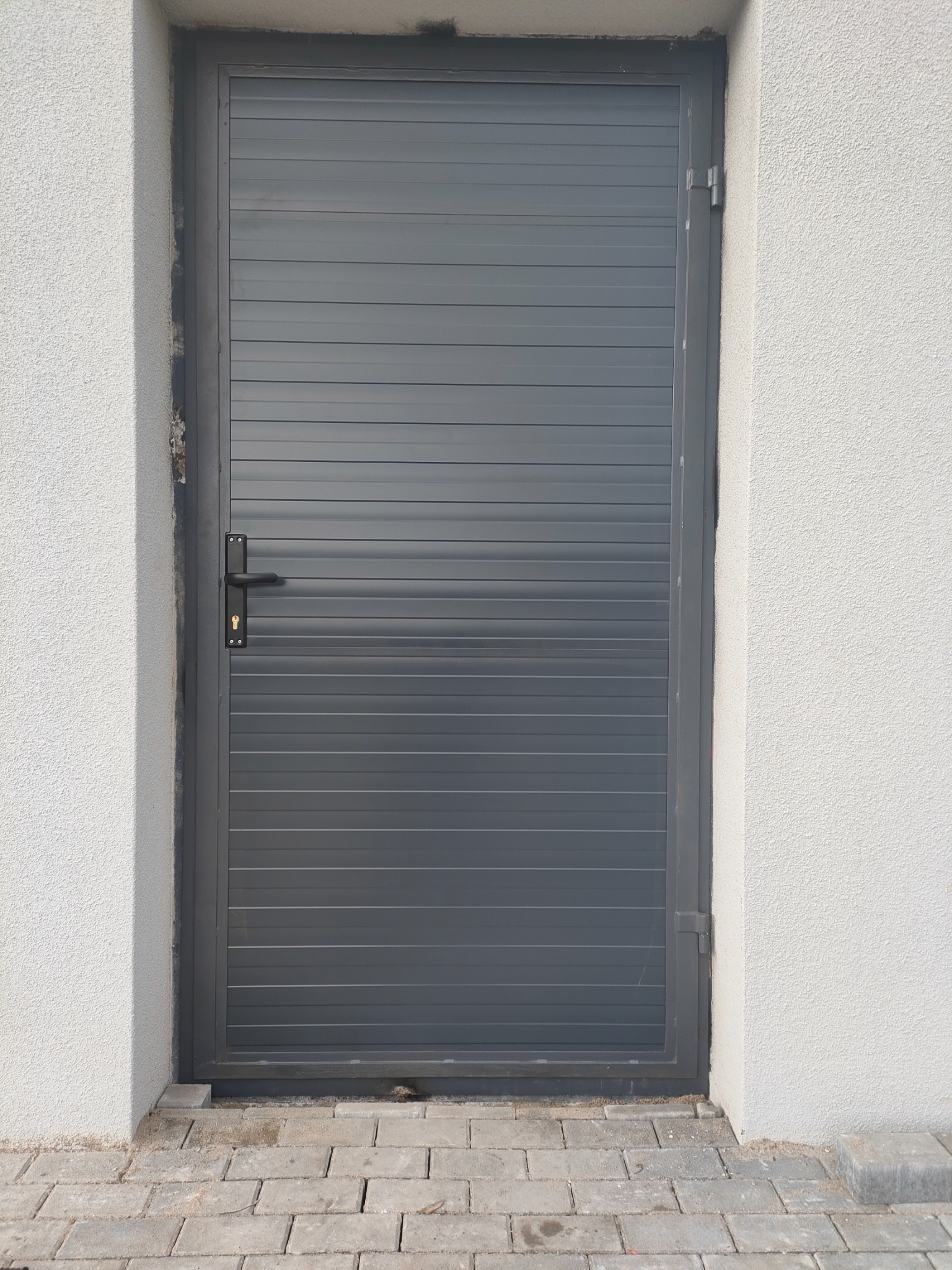 Szara brama wjazdowa z poziomymi panelami, zamontowana w białym murze z kostką brukową u podstawy.