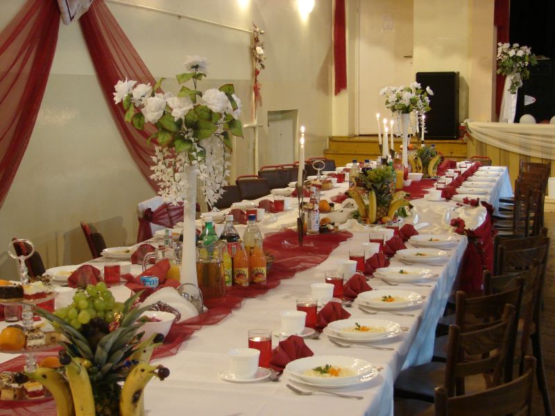 Uroczysty stół weselny z białym obrusem, czerwonym bieżnikiem i dekoracjami kwiatowymi oraz owocami, przygotowany dla gości.