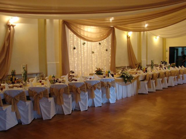 Uroczysta sala weselna z długim stołem udekorowanym białymi obrusami, kwiatami i złotymi wstążkami na krzesłach, oświetlona ciepłym światłem i ozdobiona drapowanymi tkaninami w odcieniach beżu...