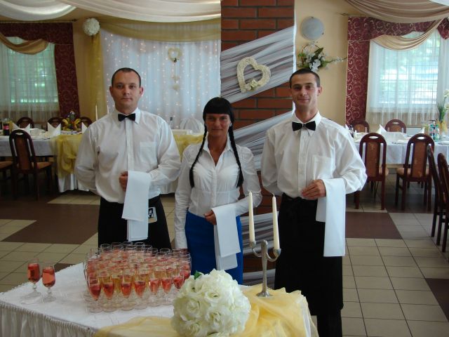 Trzyosobowy personel cateringowy w białych koszulach i czarnych muchach, prezentujący stół z piramidą kieliszków różowego szampana i bukietem białych róż na tle sali weselnej z dekoracjami.