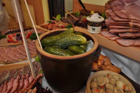 Bufet z różnorodnymi przekąskami: kiszone ogórki w glinianym garnku, wędliny, pieczarki marynowane, mięsa i inne dodatki na drewnianych deskach.
