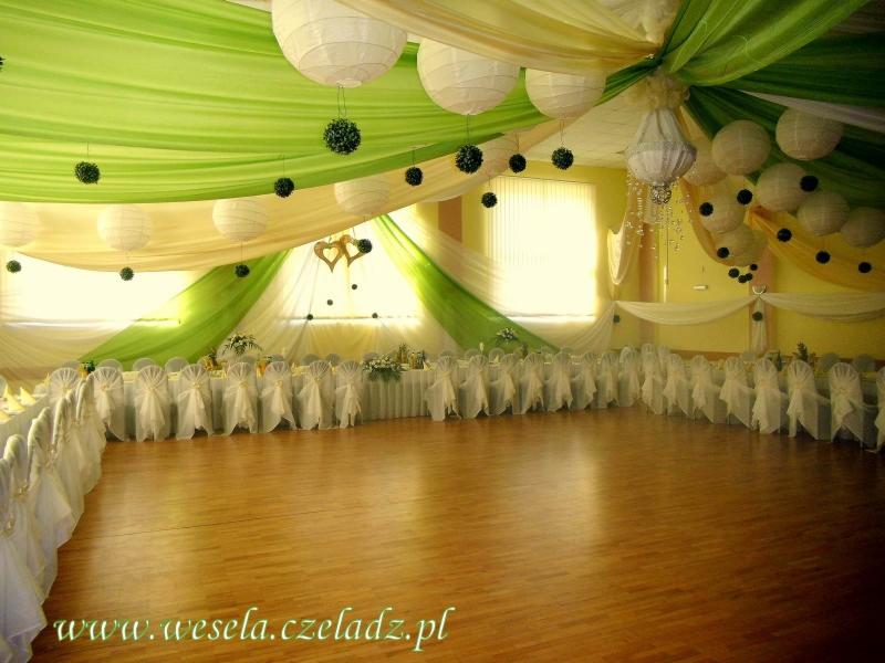 Sala balowa udekorowana zielono-żółtymi tkaninami, białymi lampionami i kulami, z długim stołem i krzesłami w białych pokrowcach.