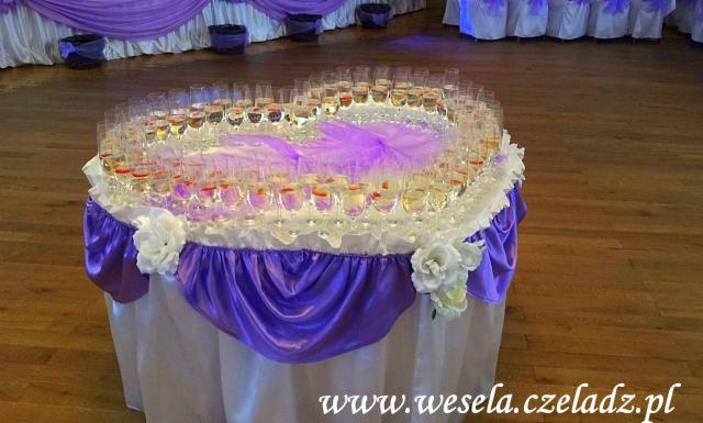 Piramida z kieliszków szampana ułożona w kształcie serca, dekoracja stołu z białym obrusem i fioletowymi draperiami oraz białymi kwiatami, w tle sala bankietowa z dekoracjami.