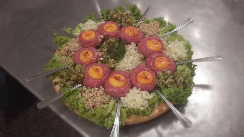 Tatar wołowy ułożony w okrąg z żółtkiem po środku, otoczony sałatą, posiekaną cebulą, ogórkiem i przyprawami, podany na drewnianej desce z wbitymi łyżeczkami.