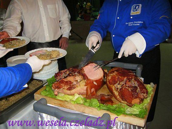 Szef kuchni w niebieskim uniformie kroi wędzoną szynkę na drewnianej desce, otoczoną sałatą, podczas obsługi gości z talerzami pełnymi jedzenia.