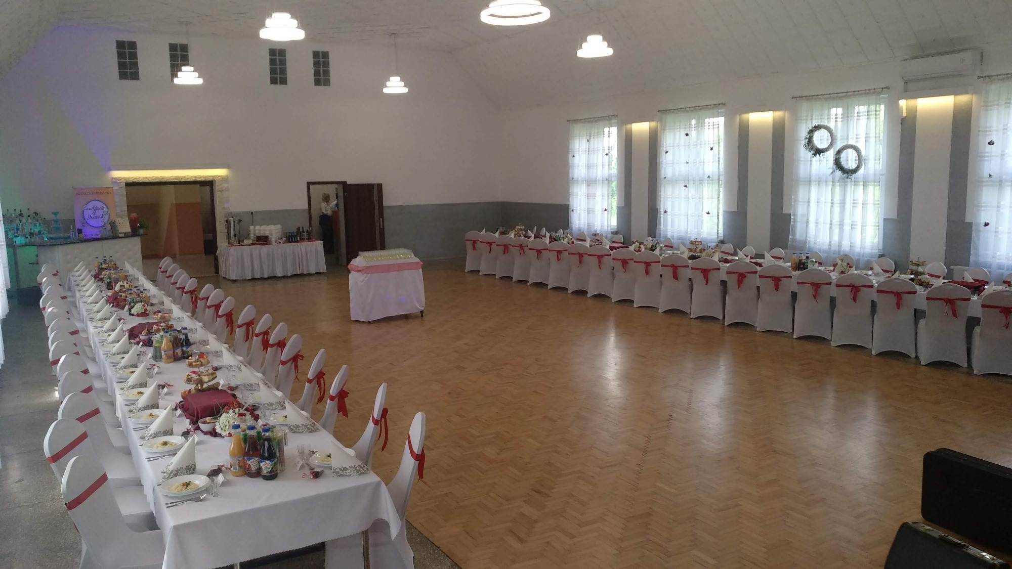 Sala bankietowa z długimi stołami udekorowanymi białymi obrusami, serwetkami i czerwonymi wstążkami na krzesłach, gotowa na przyjęcie gości, z widocznym bufetem i dekoracjami na oknach.