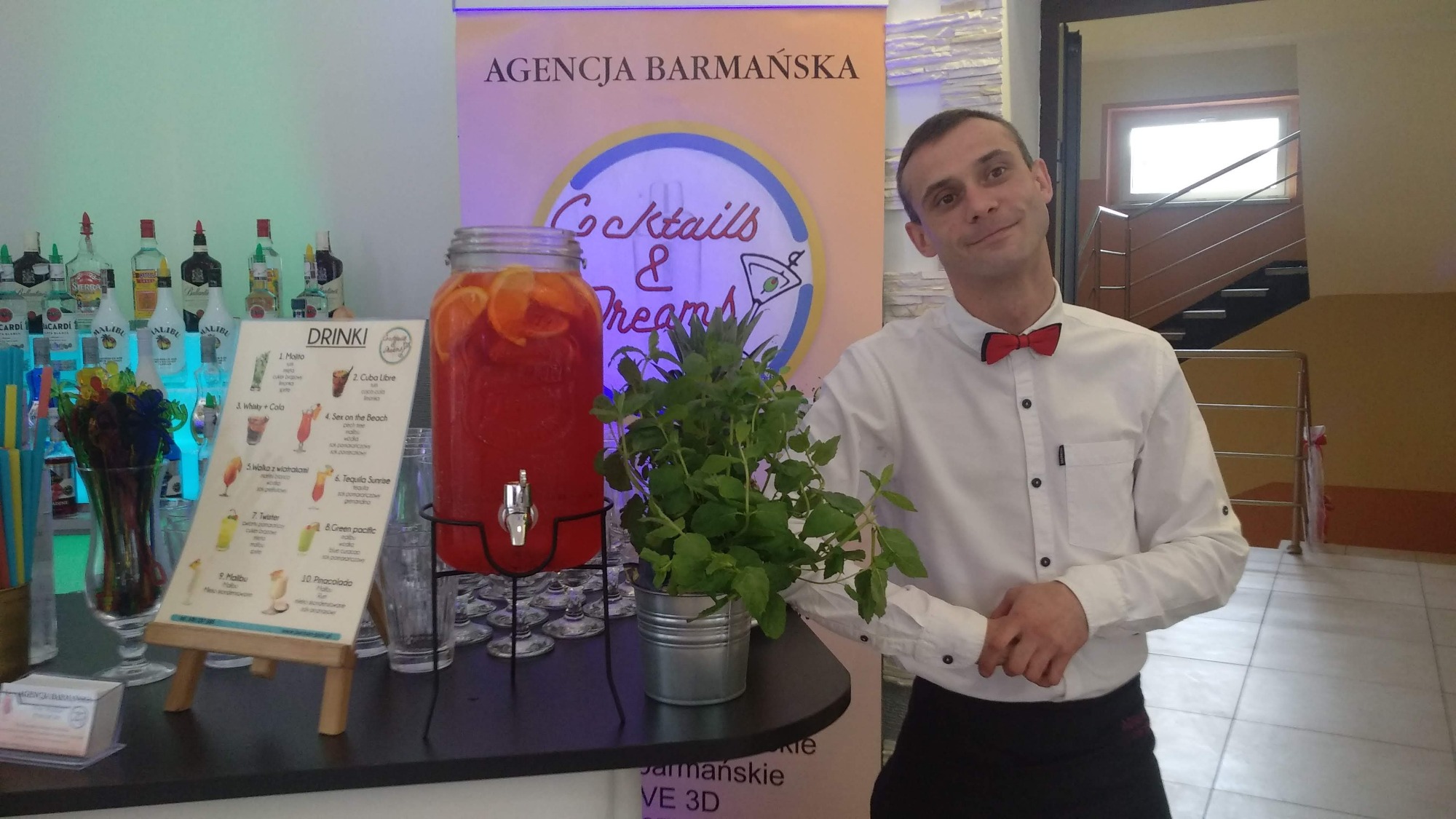 Uśmiechnięty barman w białej koszuli i czerwonej muszce stoi obok stołu z dużym słoikiem ponczu z pomarańczami, menu drinków na sztaludze i butelkami alkoholu w tle, prezentacja usług barmańskich.