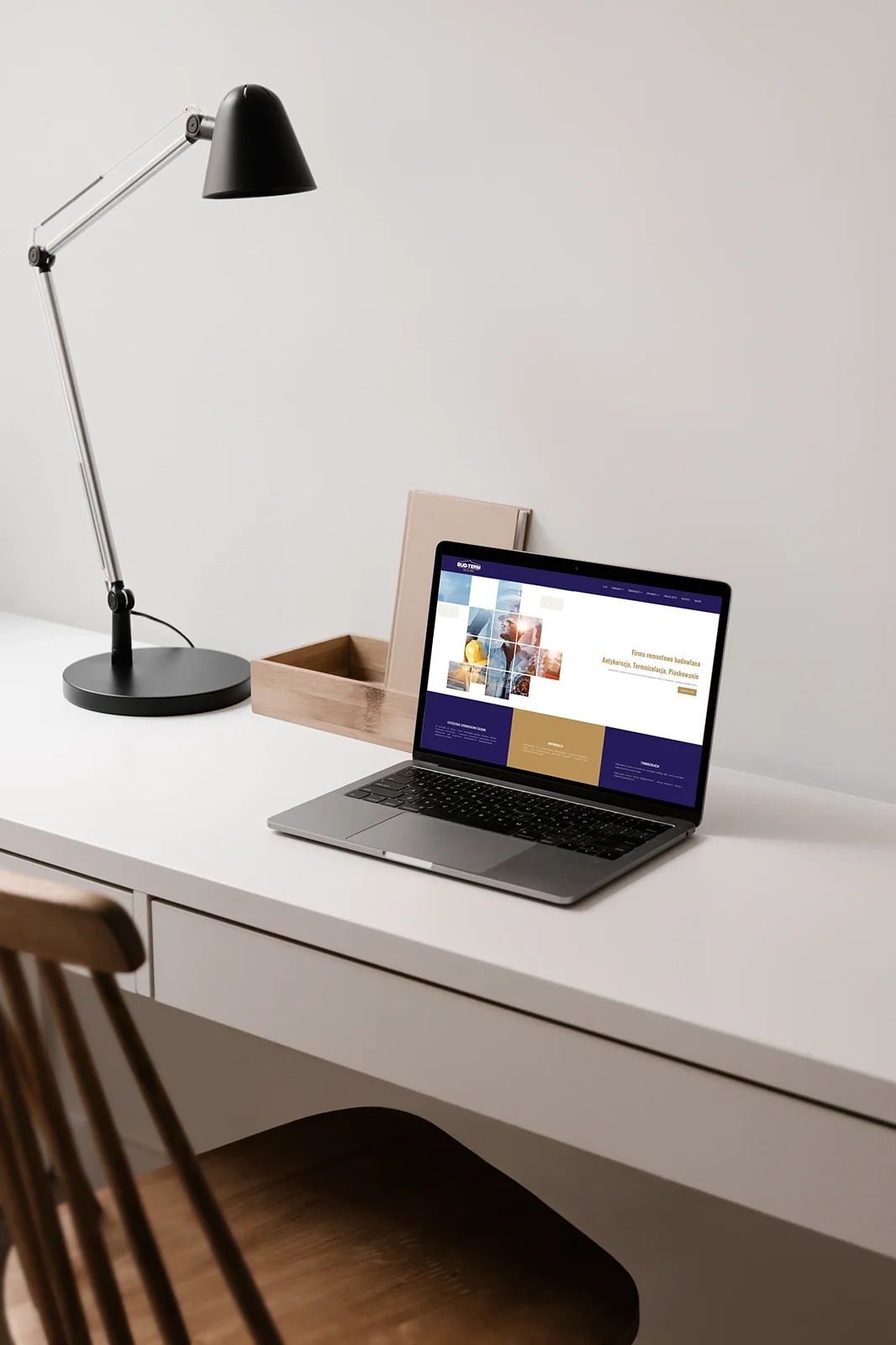 Laptop na białym biurku wyświetla stronę internetową z logo firmy budowlanej, obok lampka biurkowa i drewniane pudełko, w tle biała ściana.