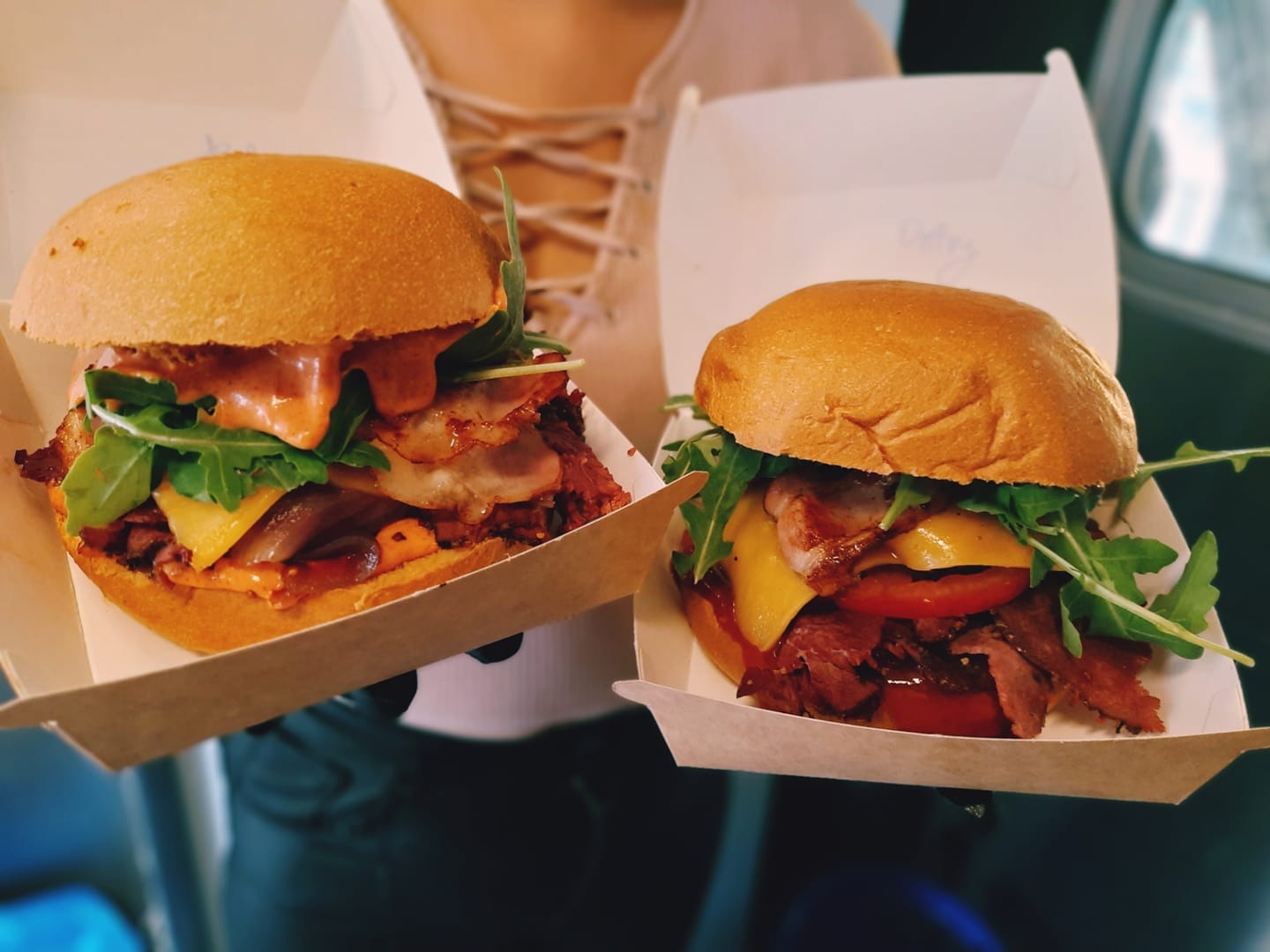 Dwa burgery w kartonowych pudełkach, jeden z sosem o konsystencji majonezu, rukolą, boczkiem, serem i karmelizowaną cebulą, drugi z rukolą, boczkiem, serem i pomidorem, trzymane przez osobę...