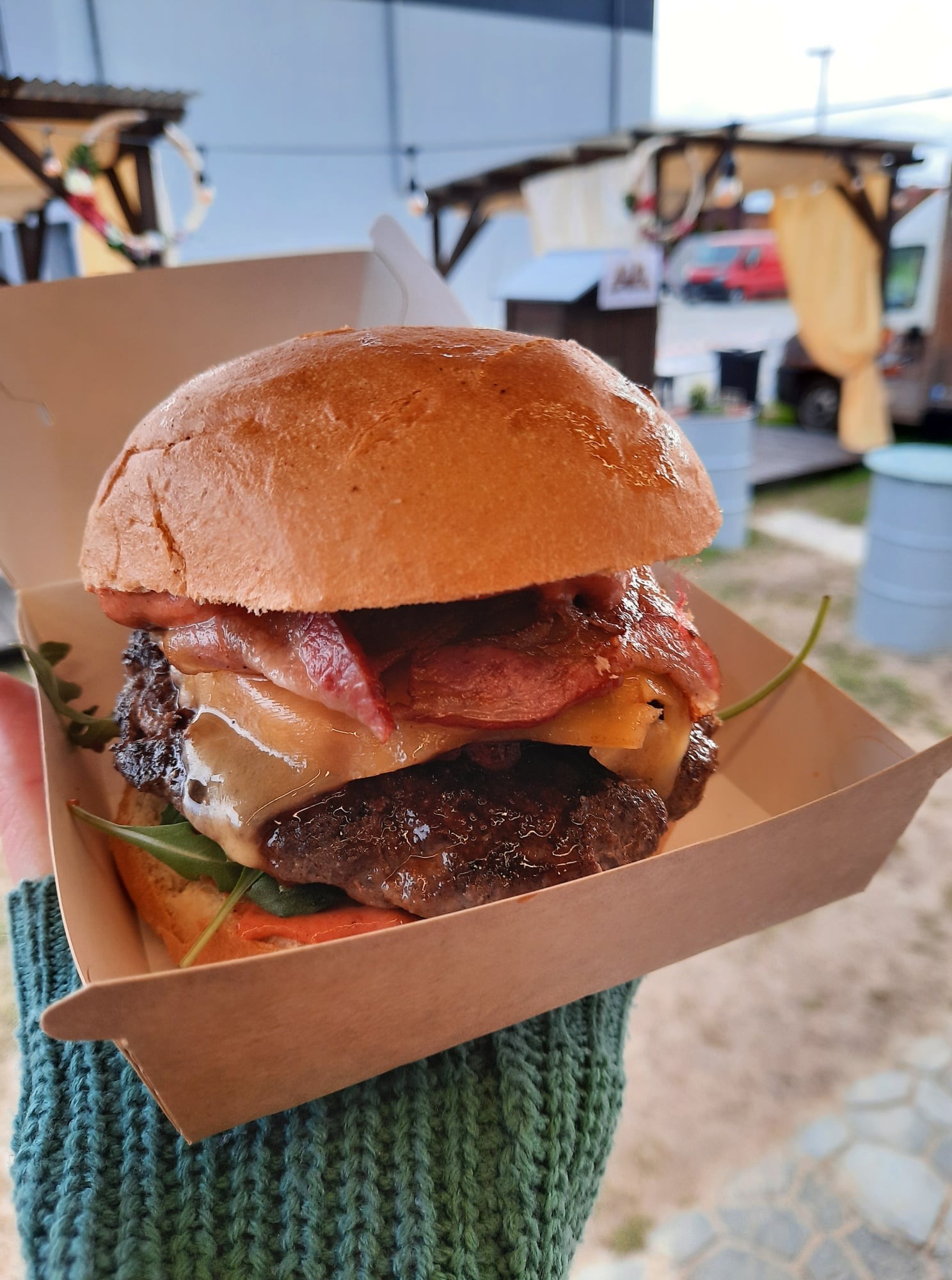 Soczysty burger z wołowiną, boczkiem i serem w kartonowym pudełku, trzymany w dłoni ubranej w zielony sweter, z rozmytym tłem plenerowej kuchni.