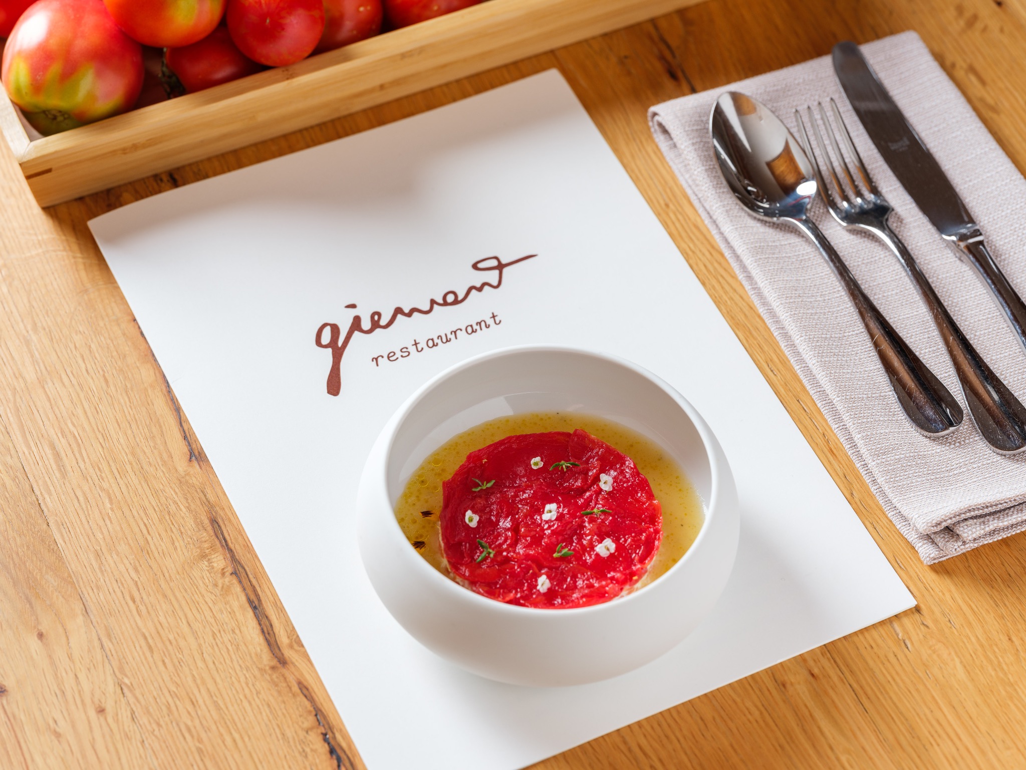 Eleganckie ujęcie dania z tartaru pomidorowego w białej miseczce, umieszczonego na karcie menu restauracji Giewont, z widocznymi pomidorami w drewnianej skrzynce w tle oraz sztućcami na lnianej...