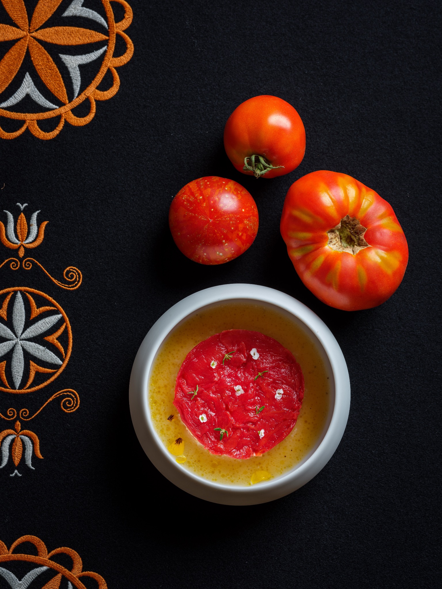 Kompozycja kulinarna: tatar z pomidorów z kwiatami i aromatyczną oliwą w białej miseczce, obok trzy całe pomidory na czarnym tle z haftowanymi ornamentami.
