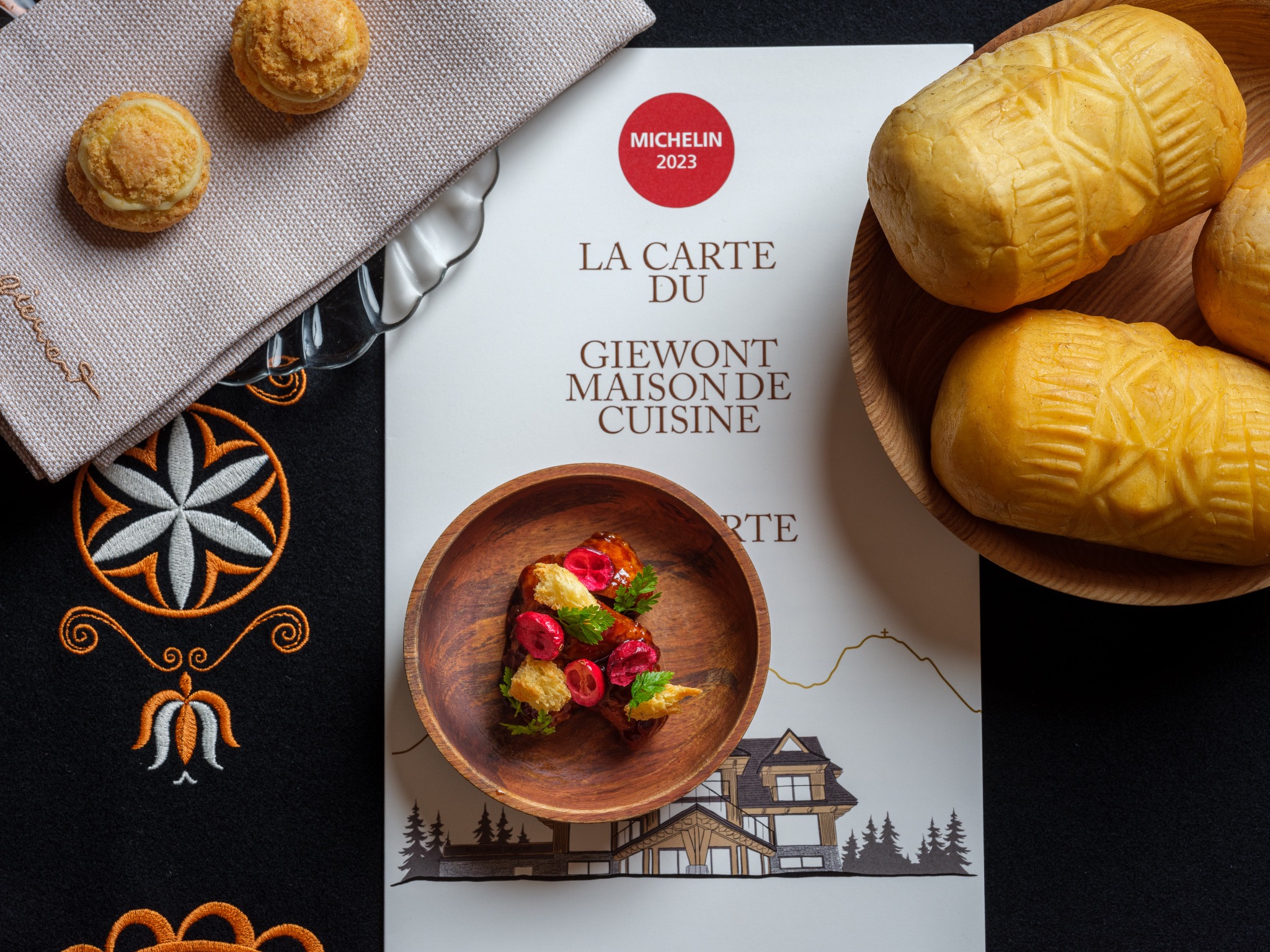 Kompozycja kulinarna: oscypki w drewnianej misce, danie z wiśniami i ziołami w mniejszej miseczce, kremowe ptysie na serwetce z haftem, karta menu restauracji Giewont z rekomendacją Michelin 2023...