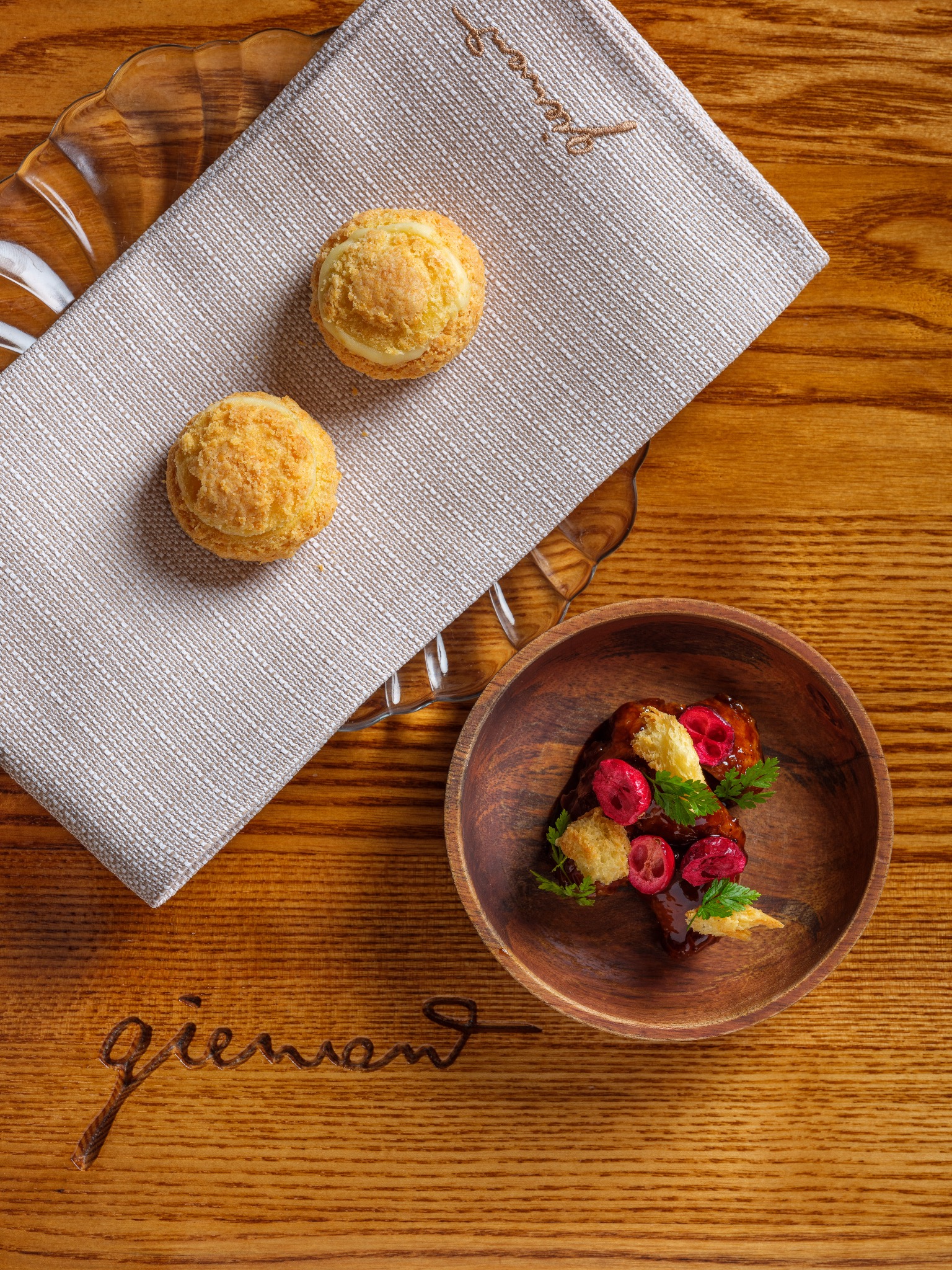 Kompozycja kulinarna z góry: dwa ptysie na lnianej serwetce z haftem, drewniana miseczka z daniem mięsnym, żurawiną i ziołami, całość na drewnianym stole z wygrawerowanym napisem 'giewont'.