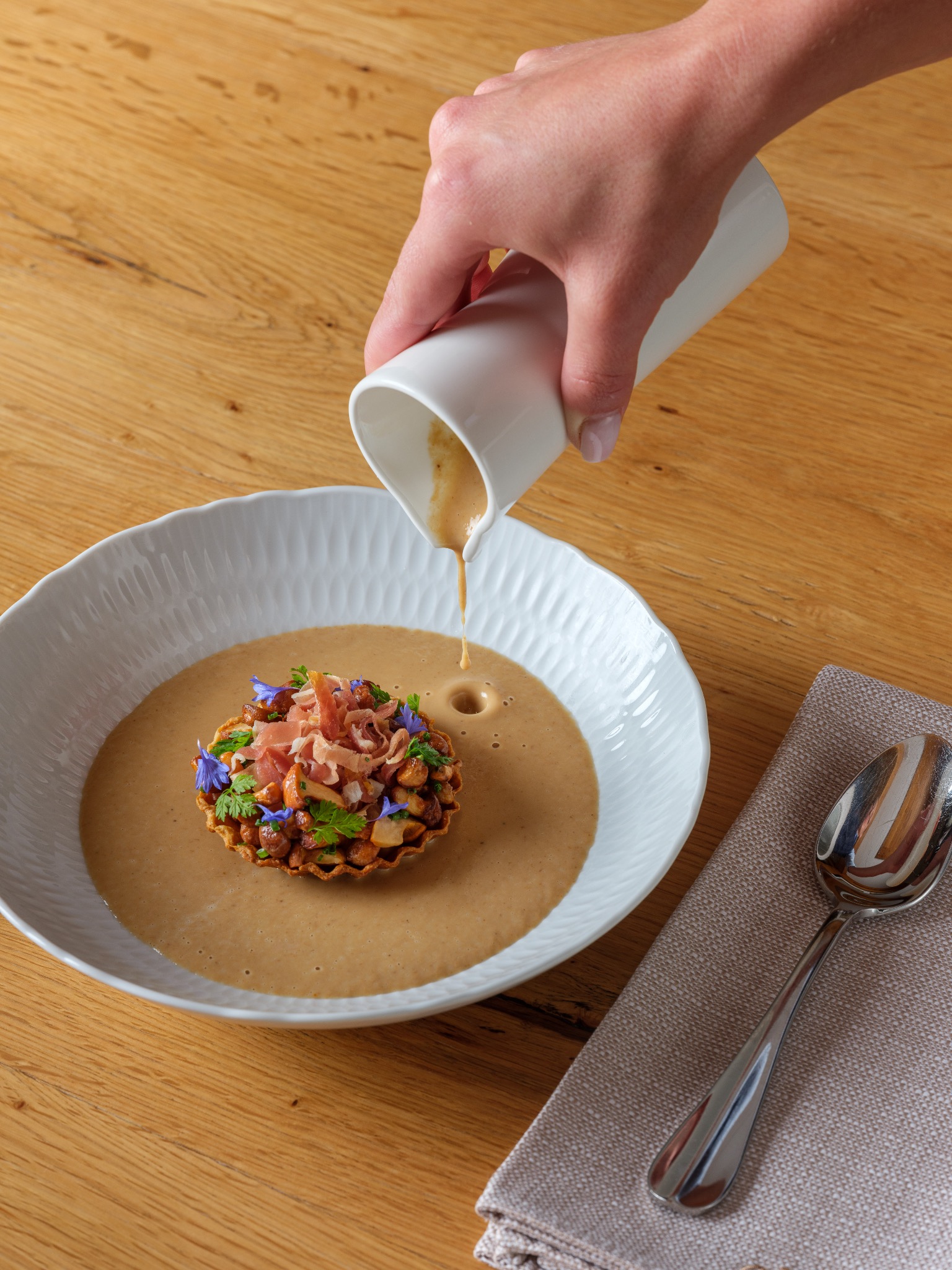 Elegancka kompozycja kulinarnej kreacji: tarta z farszem z grzybów i orzechów, dekorowana szynką parmeńską i bratkami, zalewana kremowym sosem z białego dzbanka na tle drewnianego stołu obok...