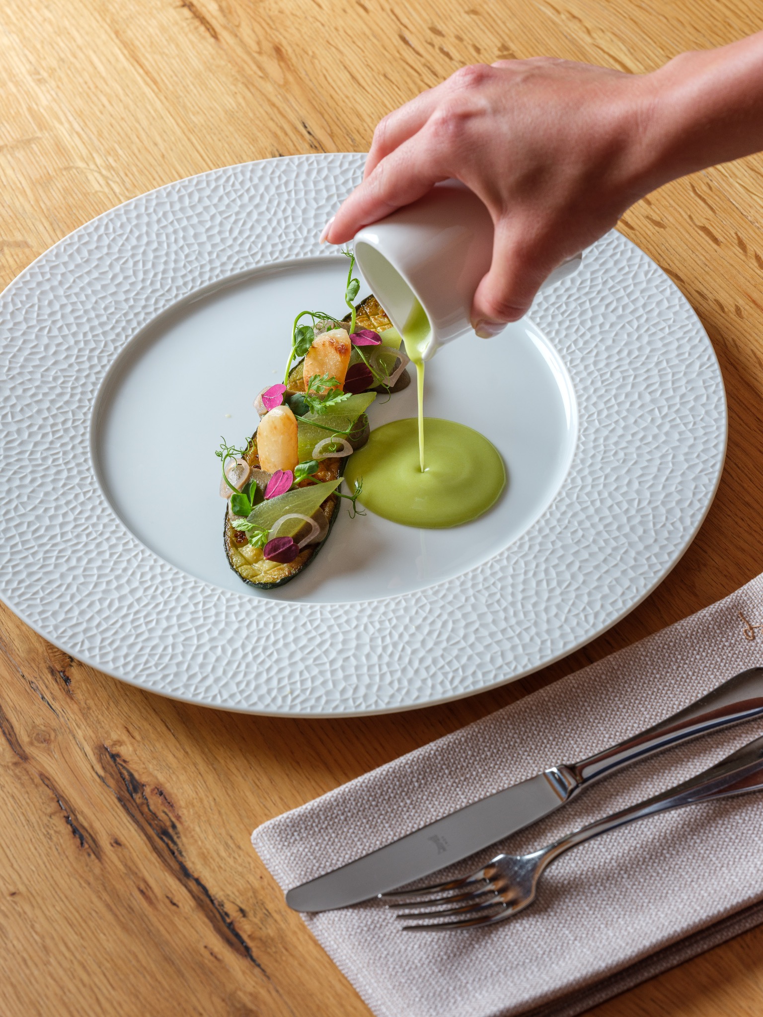 Elegancko podane danie z grillowanego bakłażana, warzyw i ziół na białym talerzu o fakturze plastra miodu, polewane zielonym sosem z małego dzbanka; obok leży sztućce na lnianej serwetce, całość...