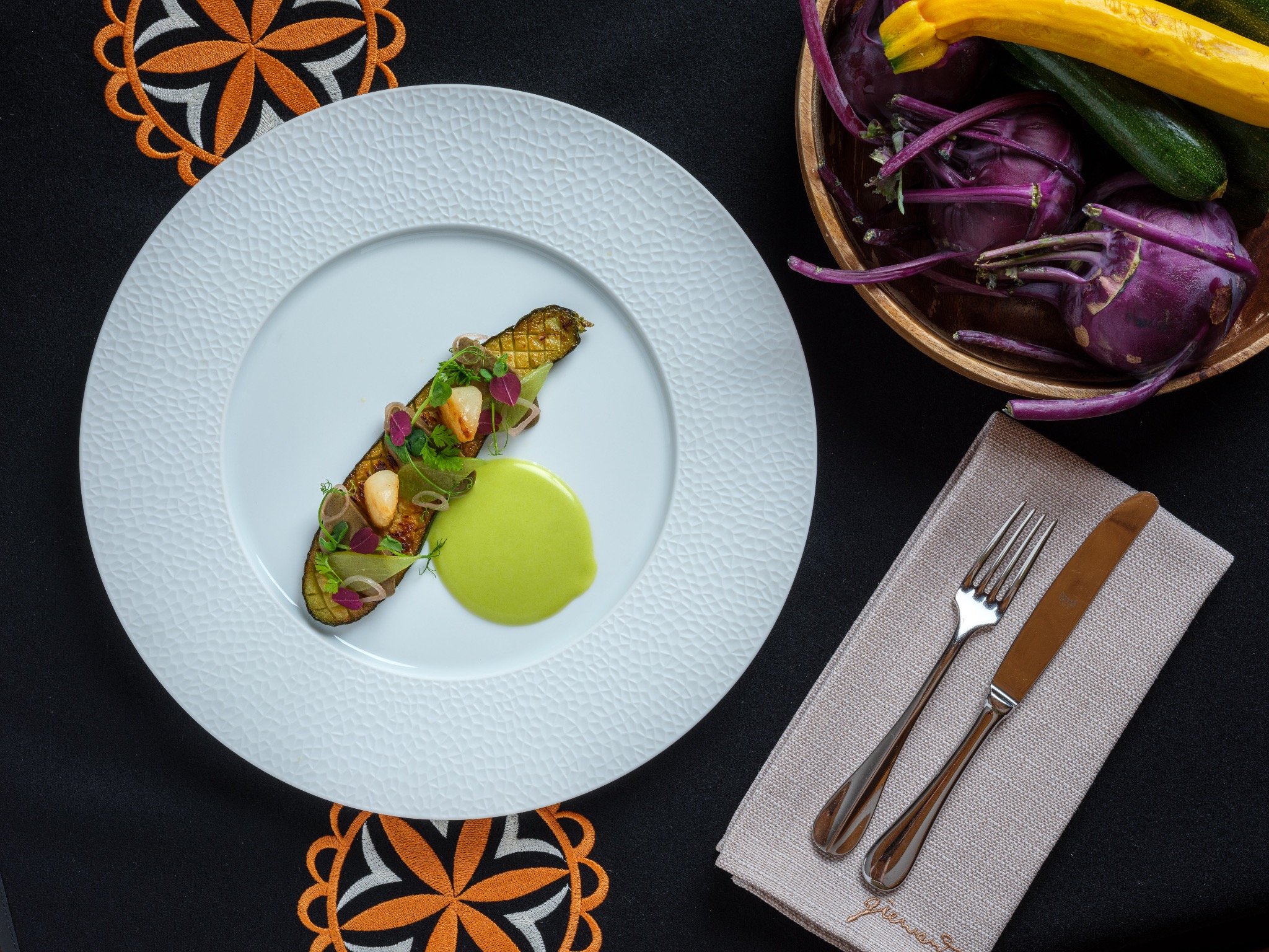 Elegancko podane danie z grillowanej cukinii z dodatkami na białym talerzu o fakturze, obok sztućce na lnianej serwetce, w tle drewniana misa z warzywami: fioletowa kalarepa, żółta i zielona...