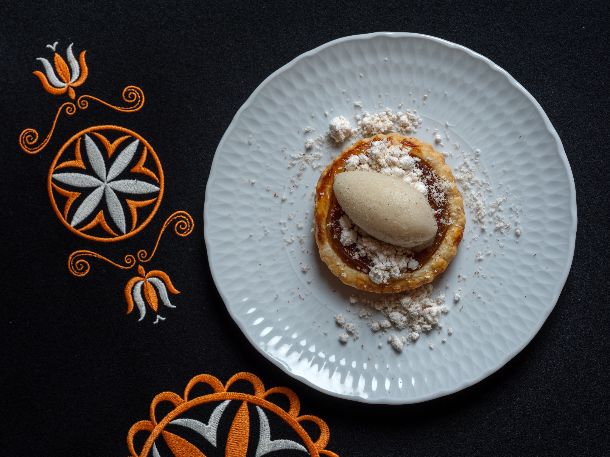 Tarta z jasnym kremem i posypką na białym talerzu o fakturze plastra miodu, na czarnym tle z pomarańczowo-białym haftem góralskim.