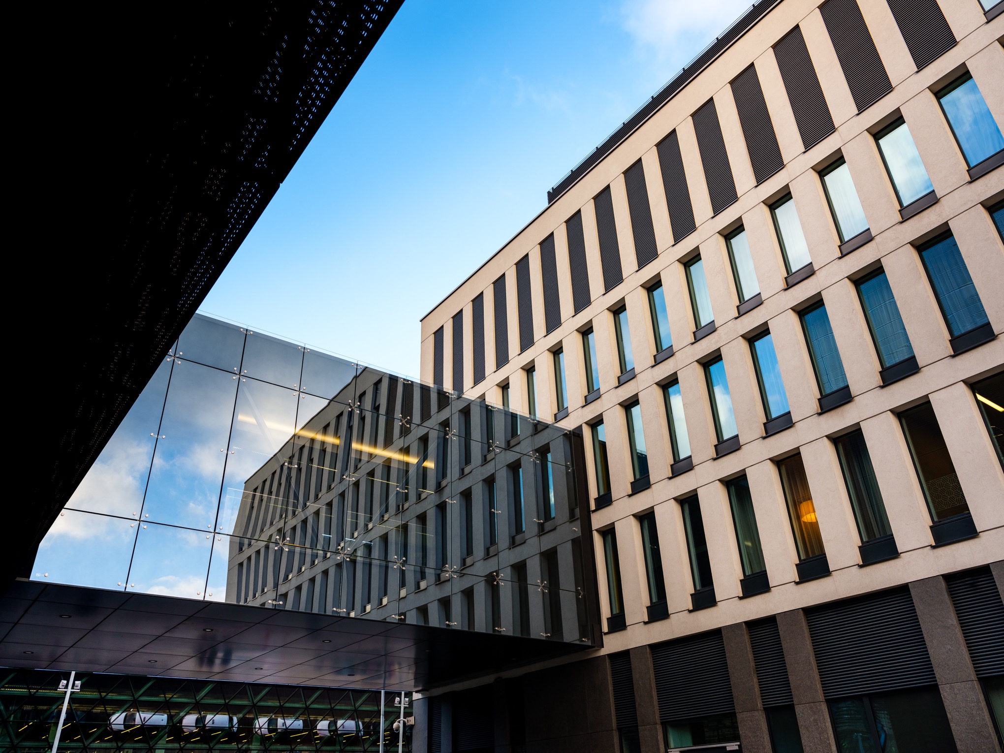 Nowoczesny budynek biurowy z przeszklonym łącznikiem, elewacja w beżowych i ciemnoszarych pasach, błękitne niebo odbijające się w szklanych powierzchniach.