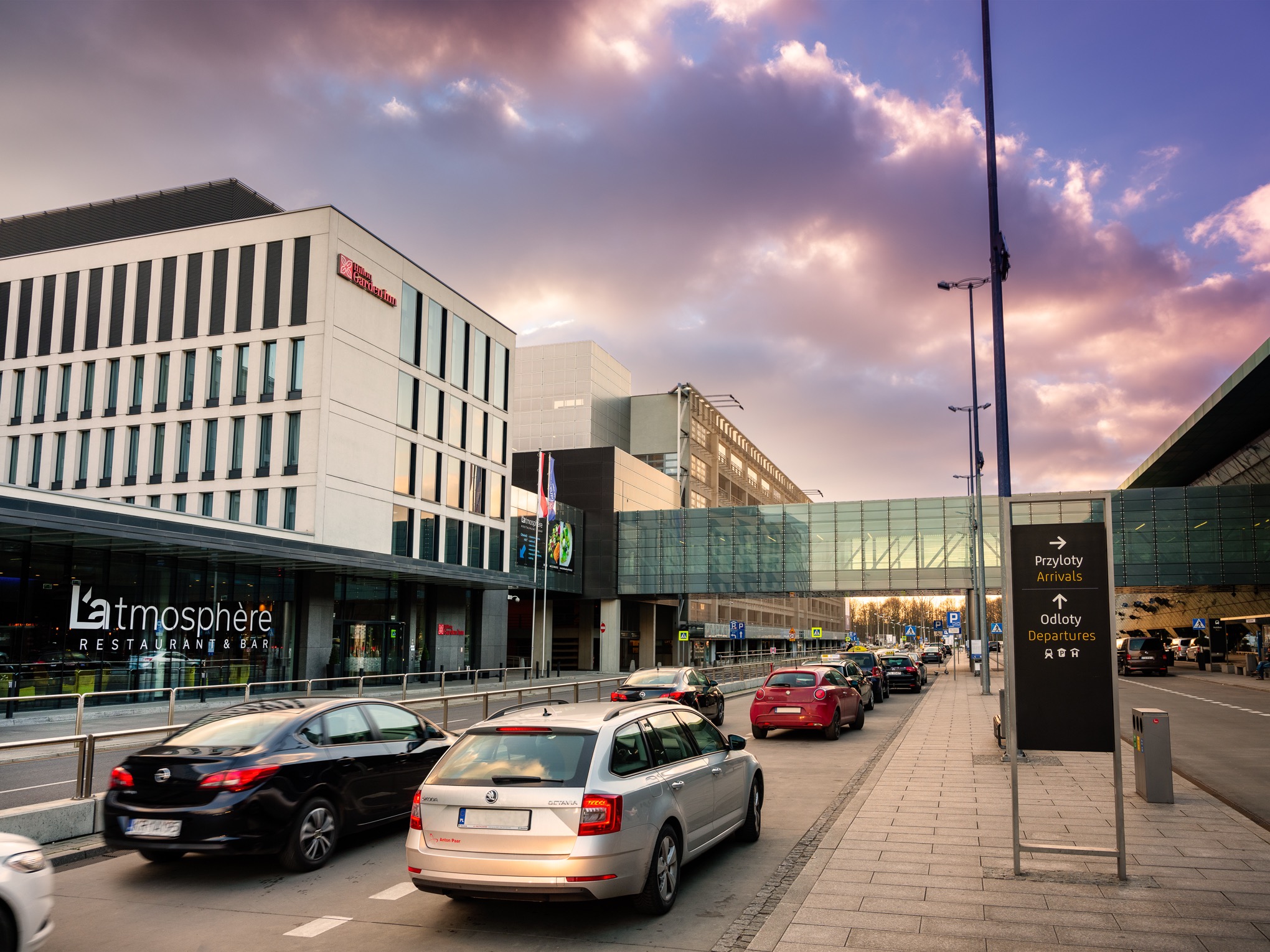 Hotel Hilton Garden Inn Airport w Krakowie Balicach widziany z zewnątrz, z zaparkowanymi samochodami i znakami informacyjnymi na pierwszym planie, sfotografowany o zmierzchu.