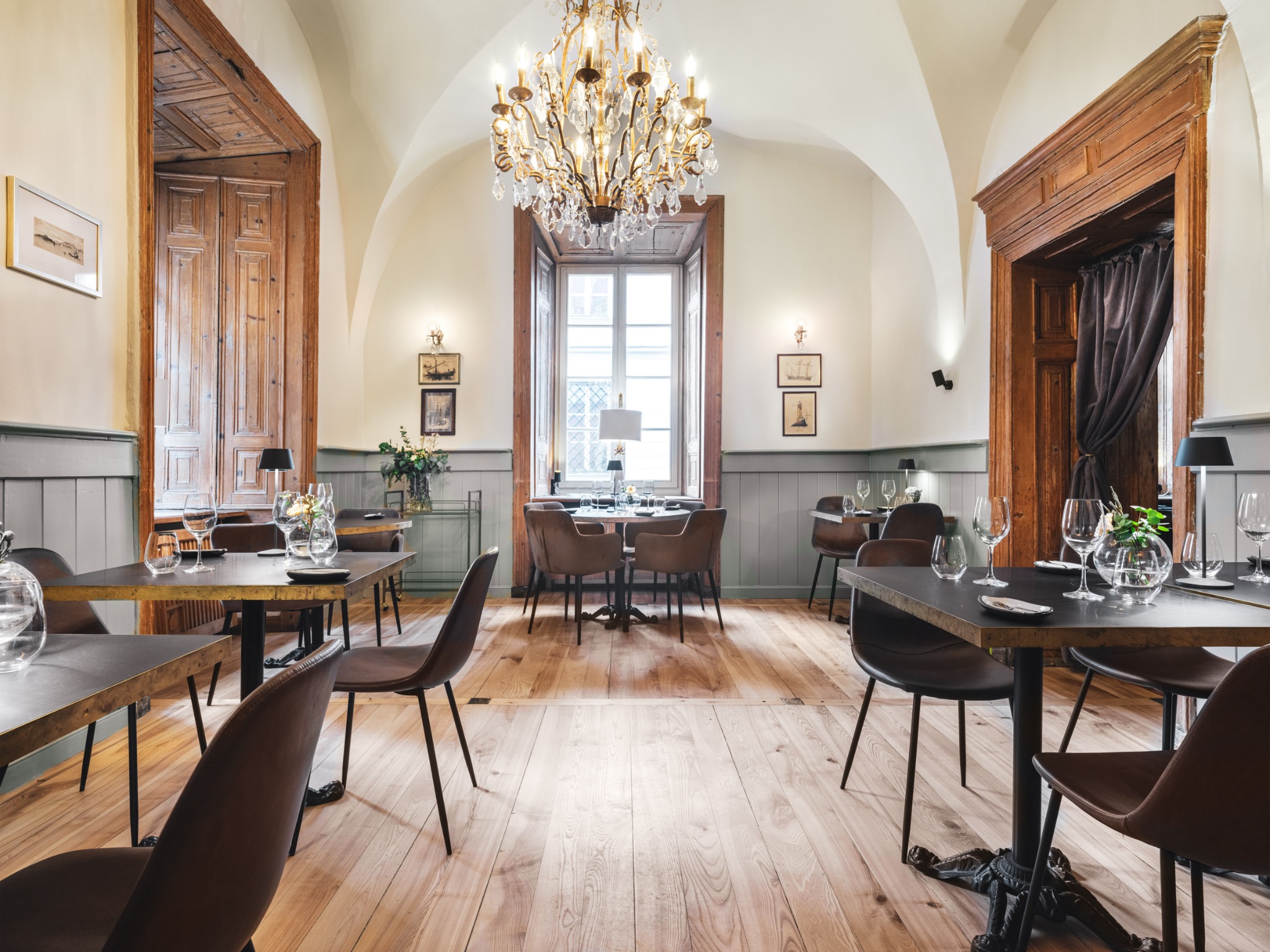 Wnętrze restauracji z drewnianą podłogą, stołami nakrytymi szkłem i sztućcami, brązowymi krzesłami, oświetlone żyrandolem z kryształkami i światłem wpadającym przez okno.