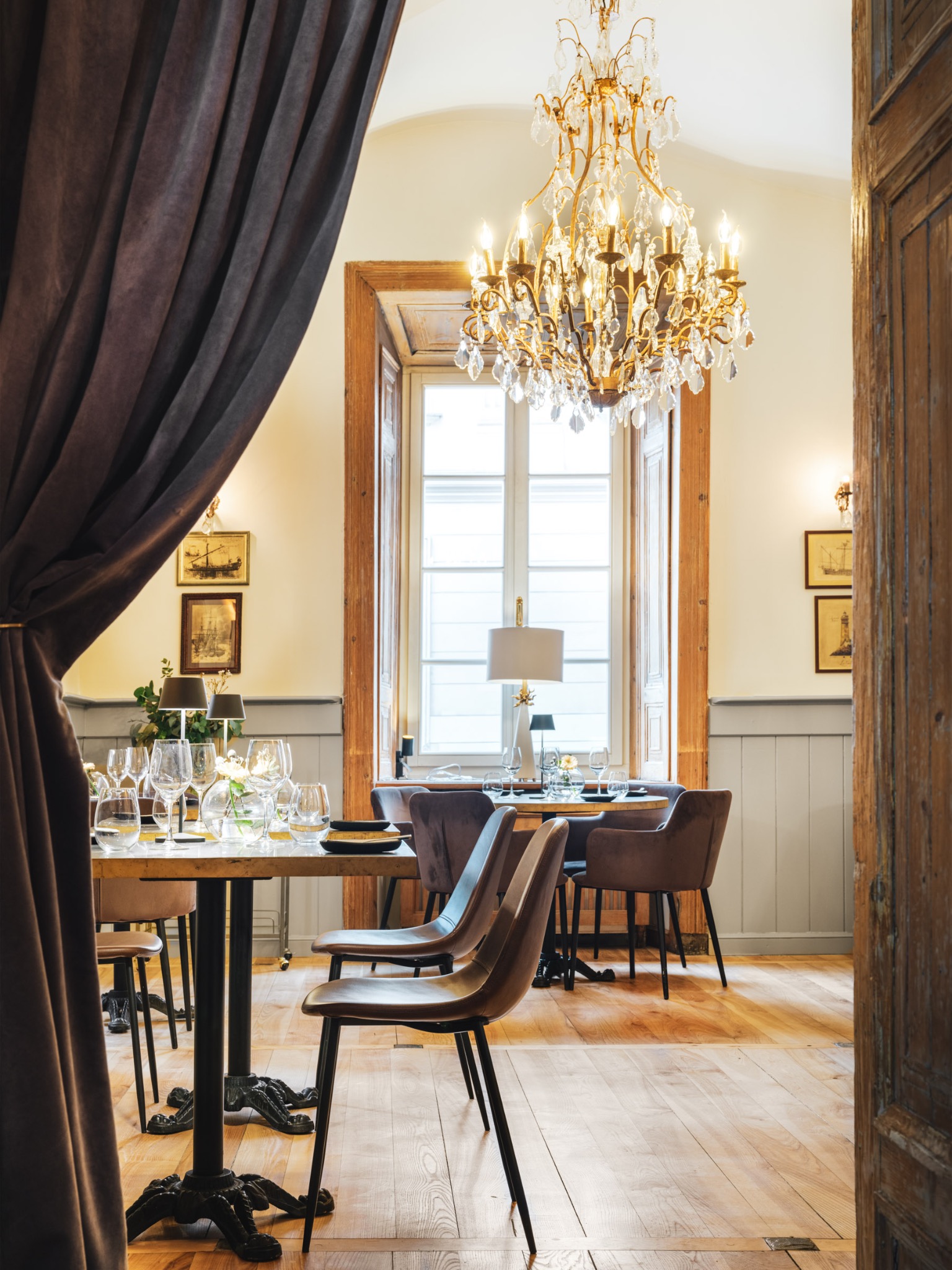Eleganckie wnętrze restauracji z drewnianą podłogą, stołami nakrytymi do posiłku, kryształowym żyrandolem i oknem z widokiem na zewnątrz, zasłonięte częściowo brązową zasłoną.