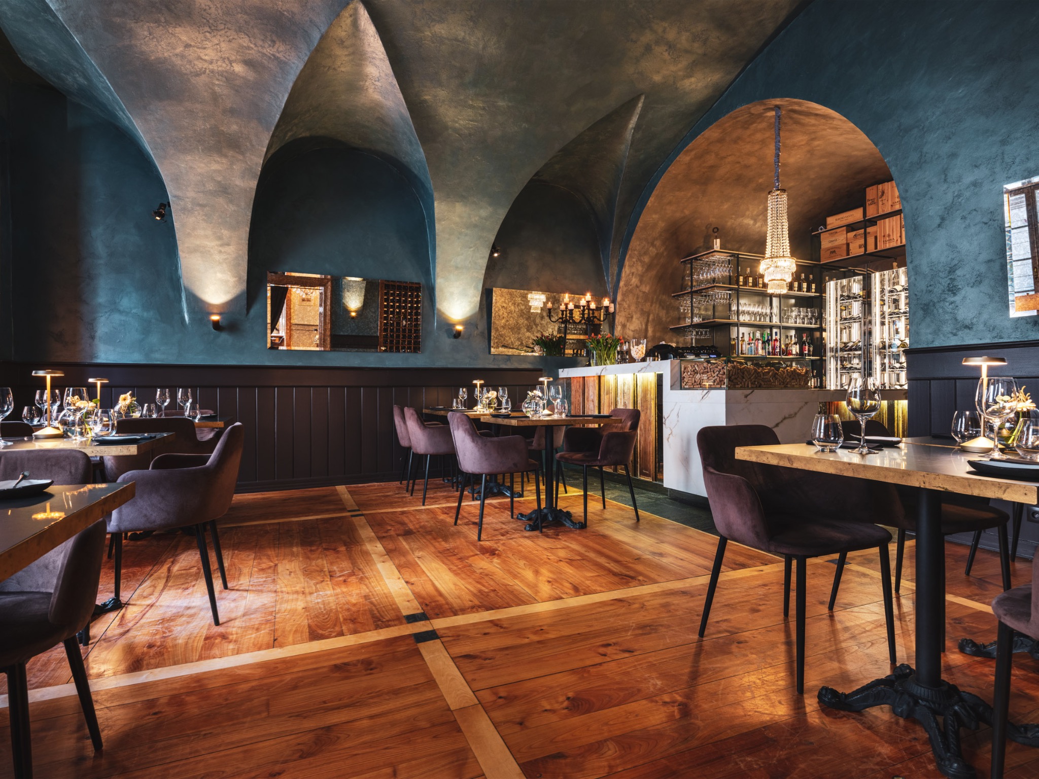 Eleganckie wnętrze restauracji: drewniana podłoga w geometryczne wzory, ciemne ściany z łukowym sufitem, stoliki z nakryciami i krzesłami w kolorze śliwki, bar z marmurowym blatem i kryształowym...