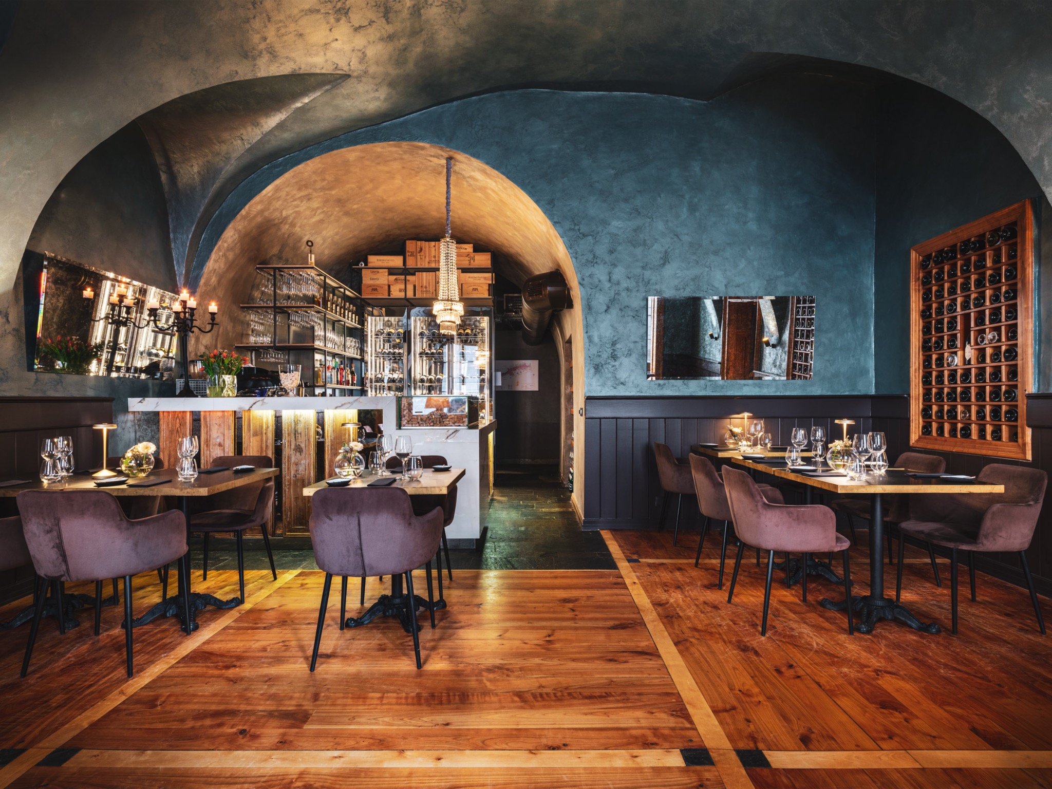 Wnętrze restauracji z drewnianą podłogą, ciemnymi ścianami i łukowym sufitem. Ustawione stoły z nakryciami i krzesłami z brązowego weluru. W tle bar z ekspozycją alkoholi i dekoracyjnymi skrzynkami.