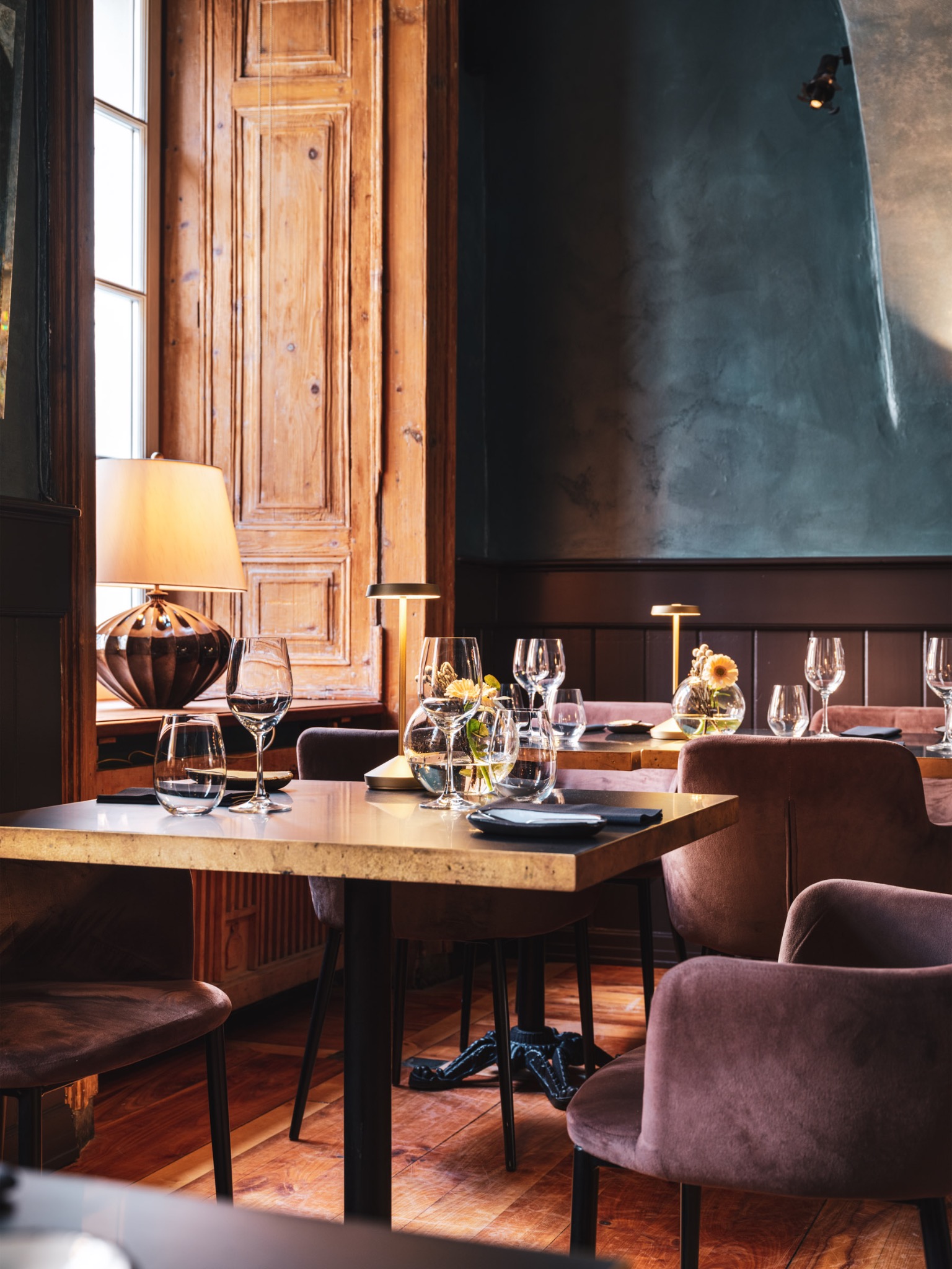 Eleganckie wnętrze restauracji z drewnianą podłogą, stołami nakrytymi szklankami i kwiatami, oświetlone lampami stołowymi i punktowym światłem na ciemnej ścianie.
